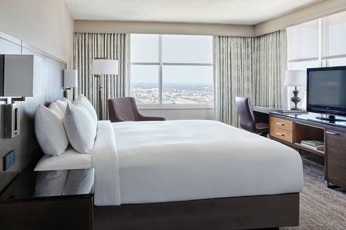 A hotel room with a large bed, a desk with a computer, a TV, and a view of the city through the window.