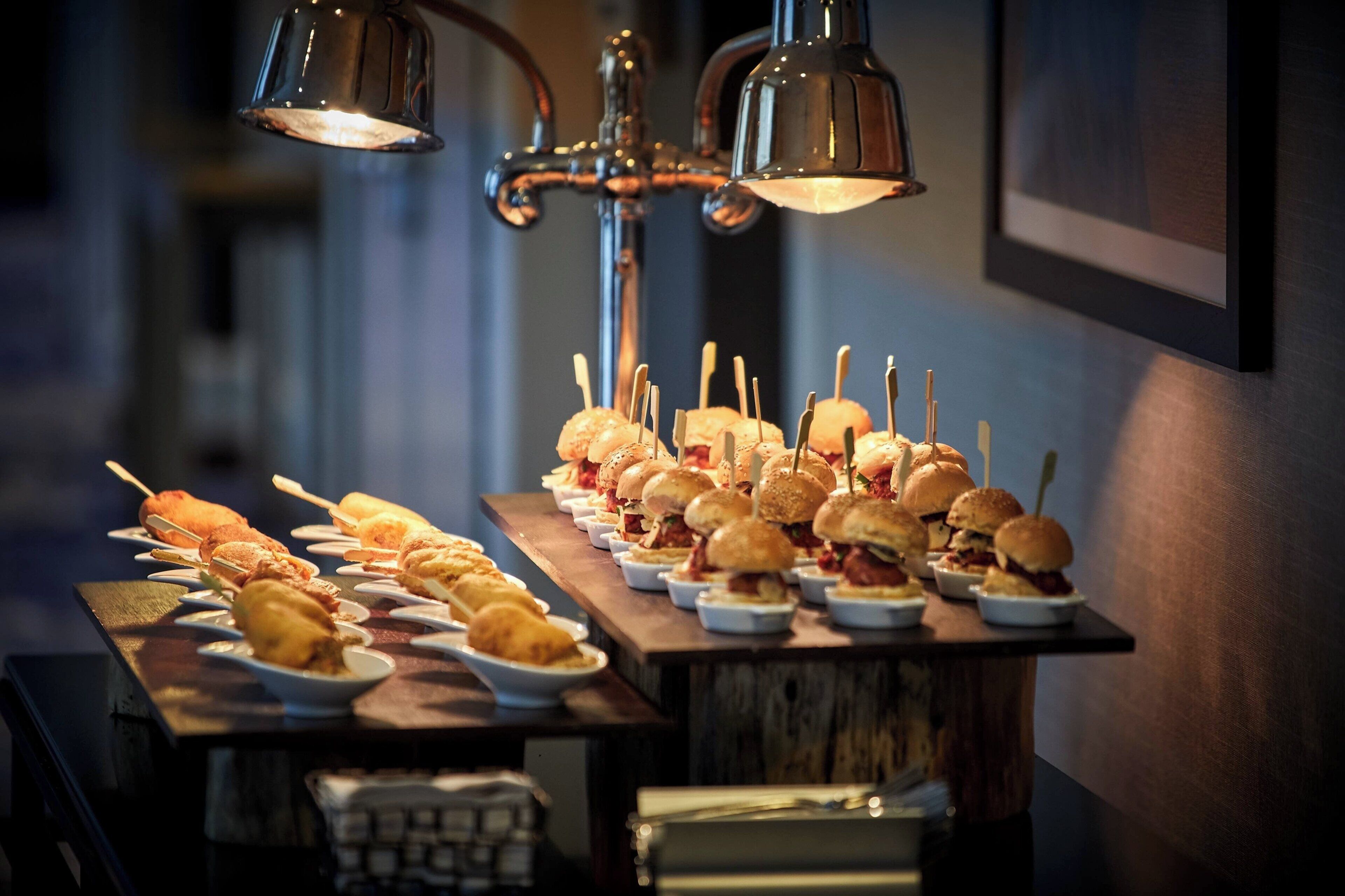 A buffet table with various dishes, including skewered items and burgers, under pendant lighting.