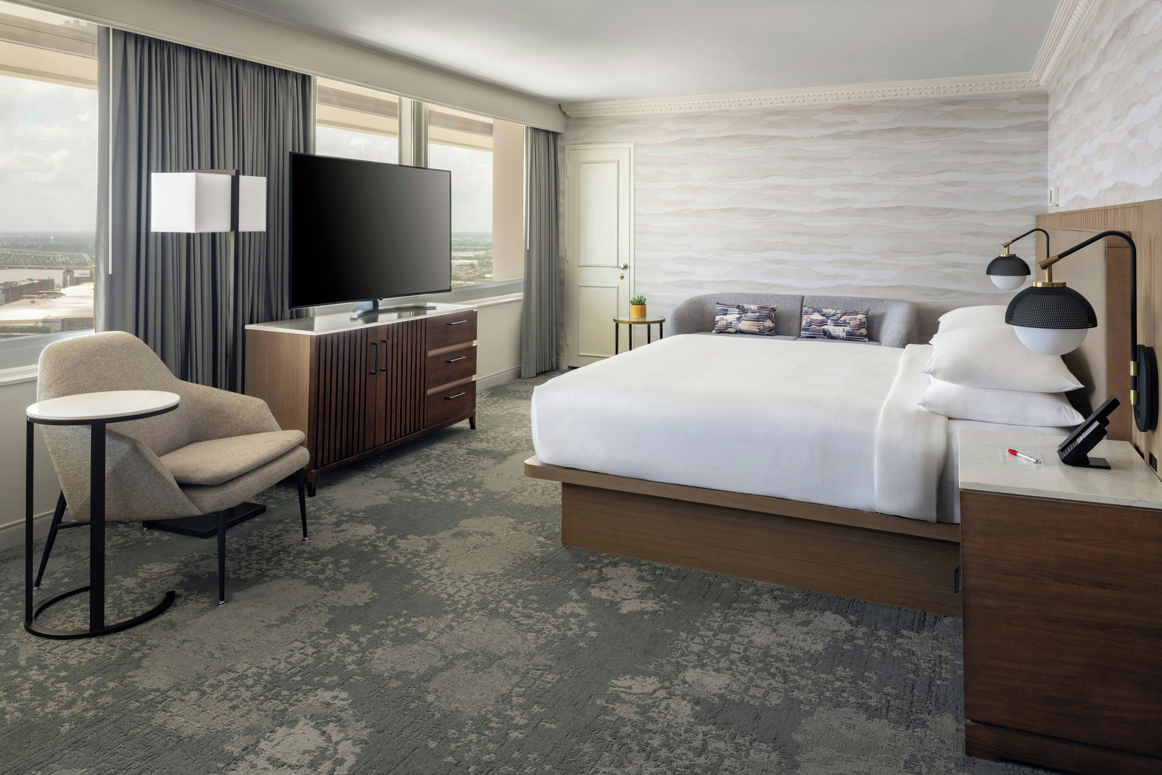 A hotel room with a bed, a TV, a chair, a desk, and a view of the city.
