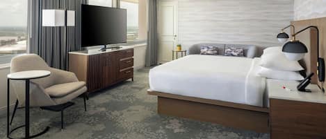 A hotel room with a bed, a TV, a chair, a desk, and a view of the city.