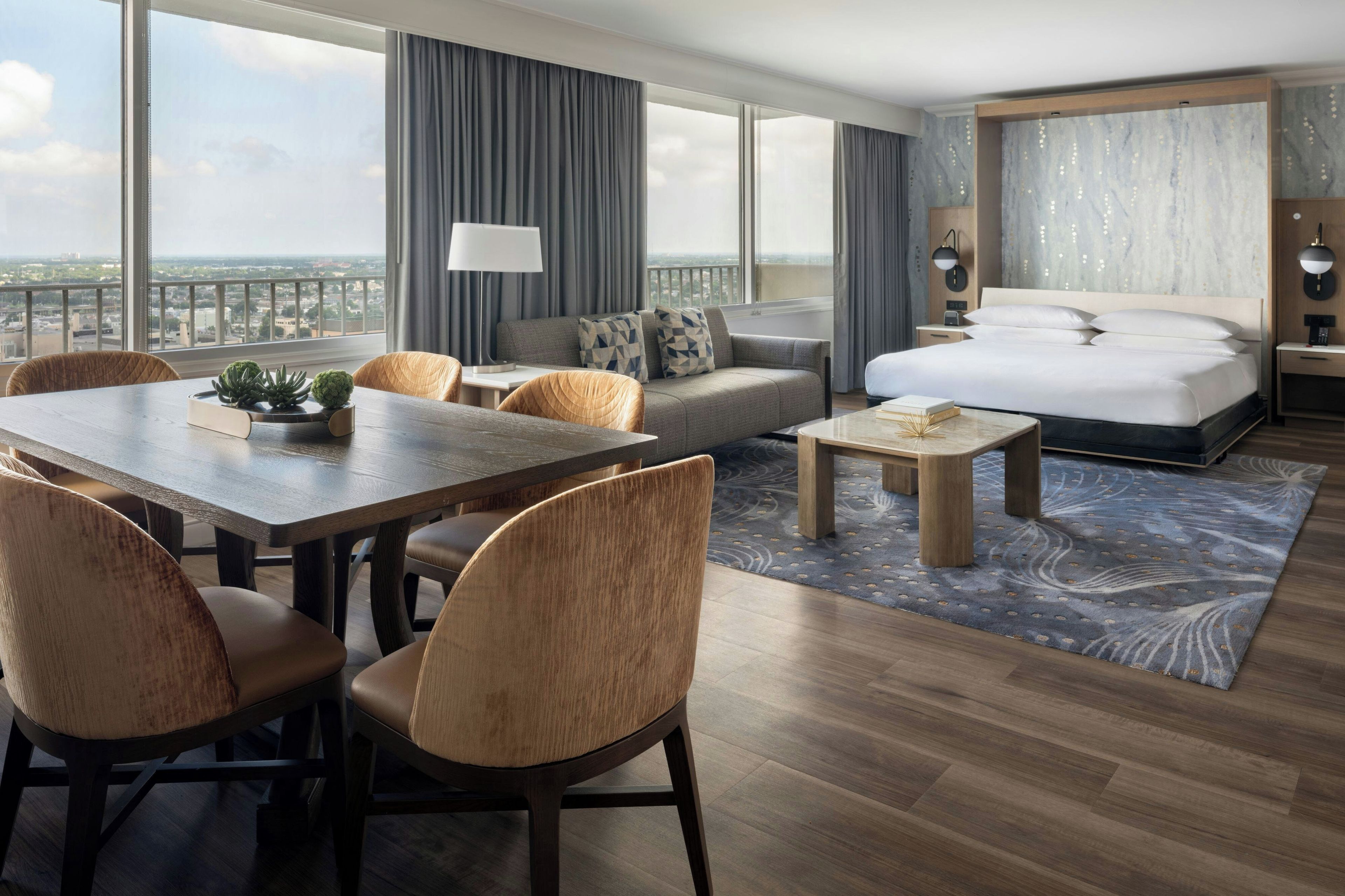Habitación de hotel moderna con una cama grande, un sofá, una mesa de comedor y sillas. Se ve la ciudad desde las ventanas.