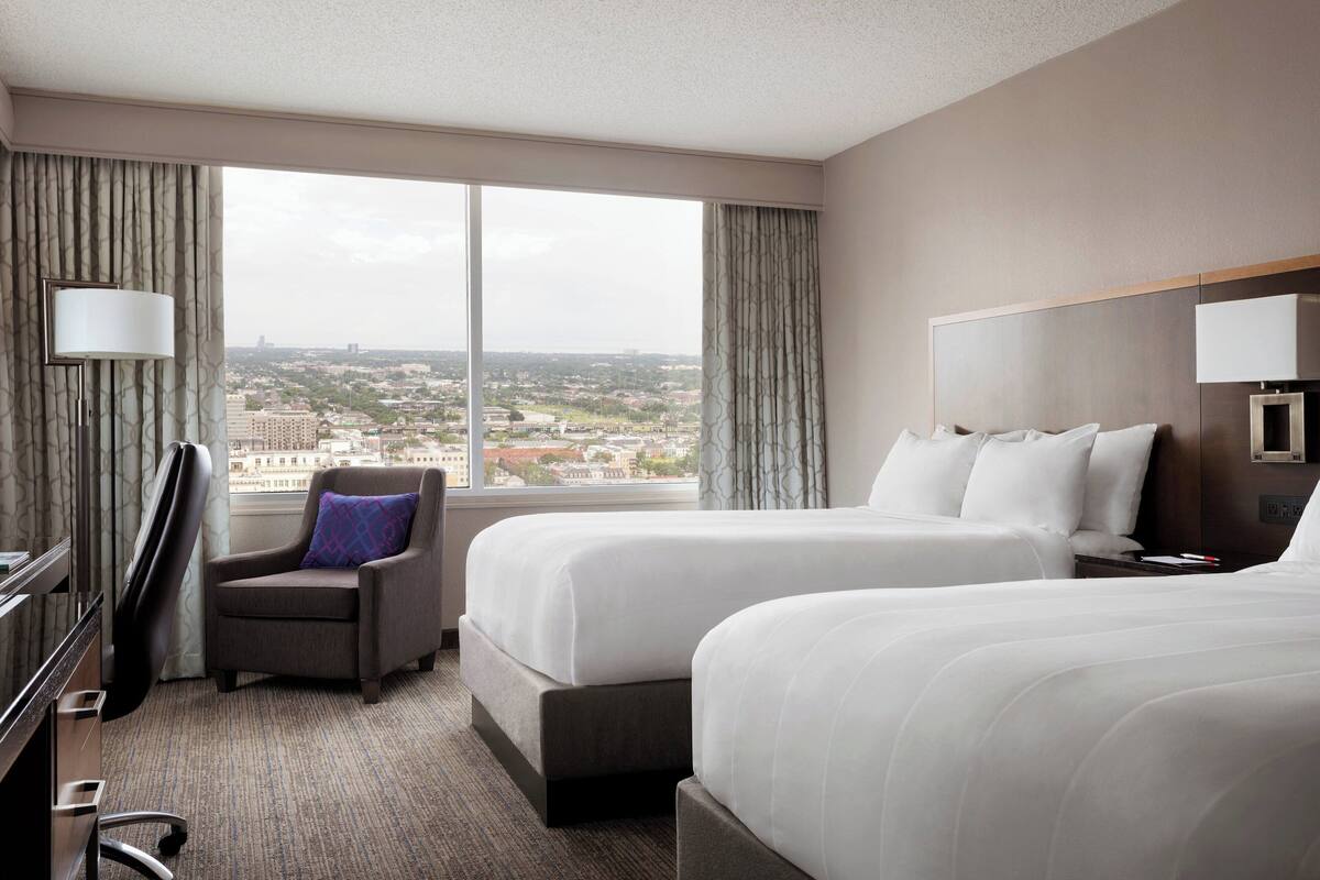 A hotel room with two beds, a desk, a chair, and a large window offering a city view.