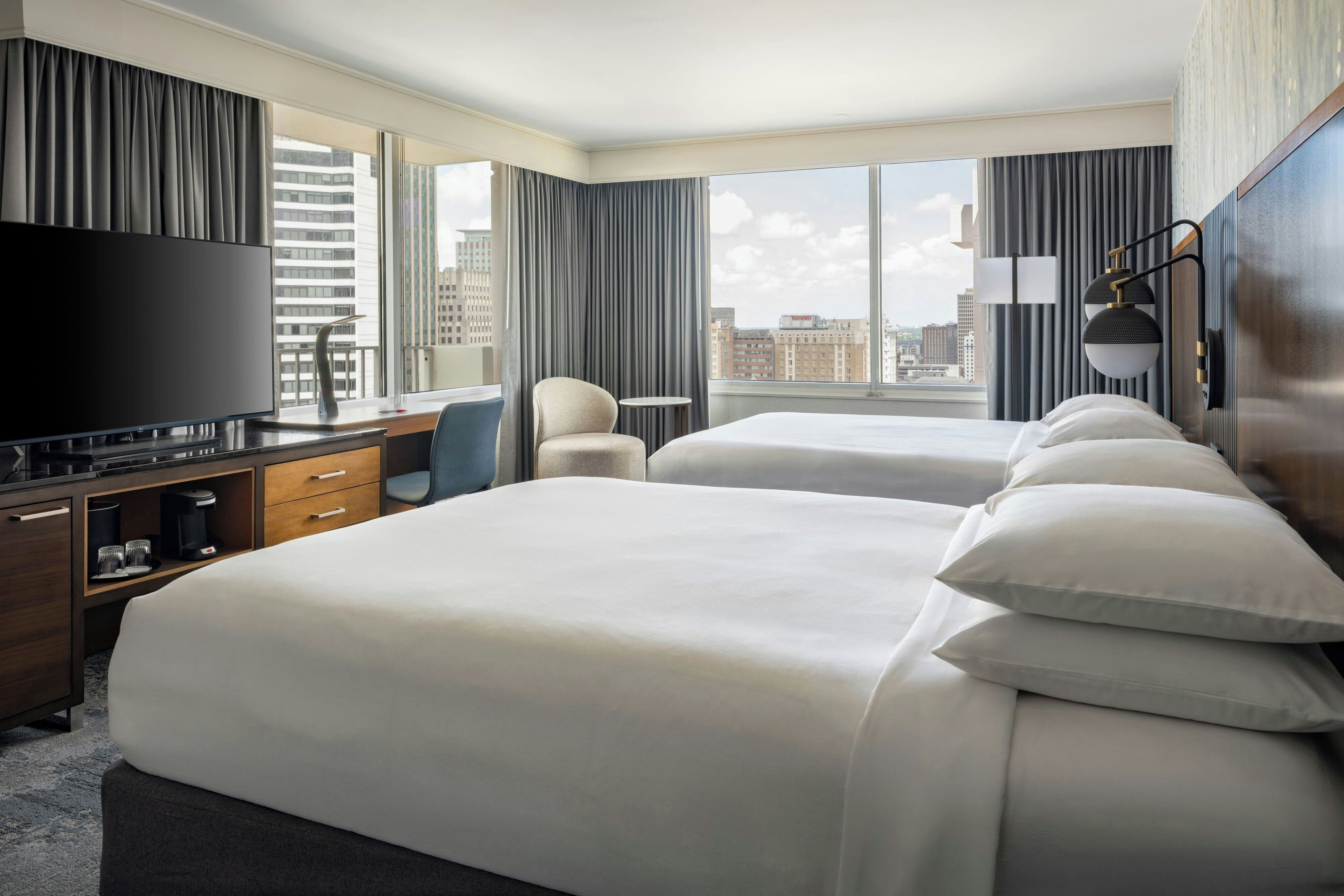 Ein Hotelzimmer mit einem großen Bett, einem Schreibtisch mit Flachbildfernseher, einem Sessel und Blick auf die Stadt durch große Fenster.