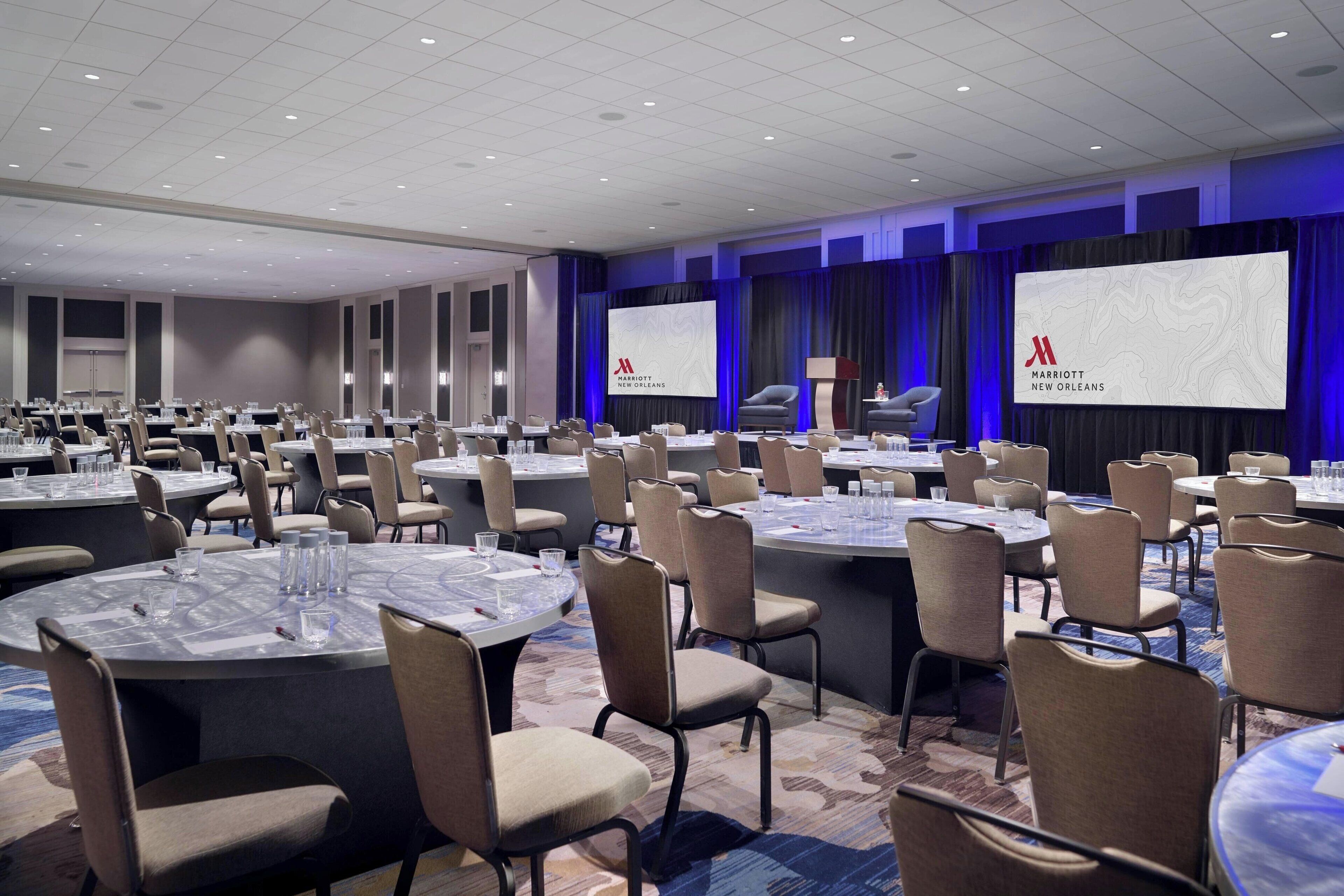 A conference room with multiple round tables and chairs, a large screen displaying the Marriott New Orleans logo, and a stage with curtains.