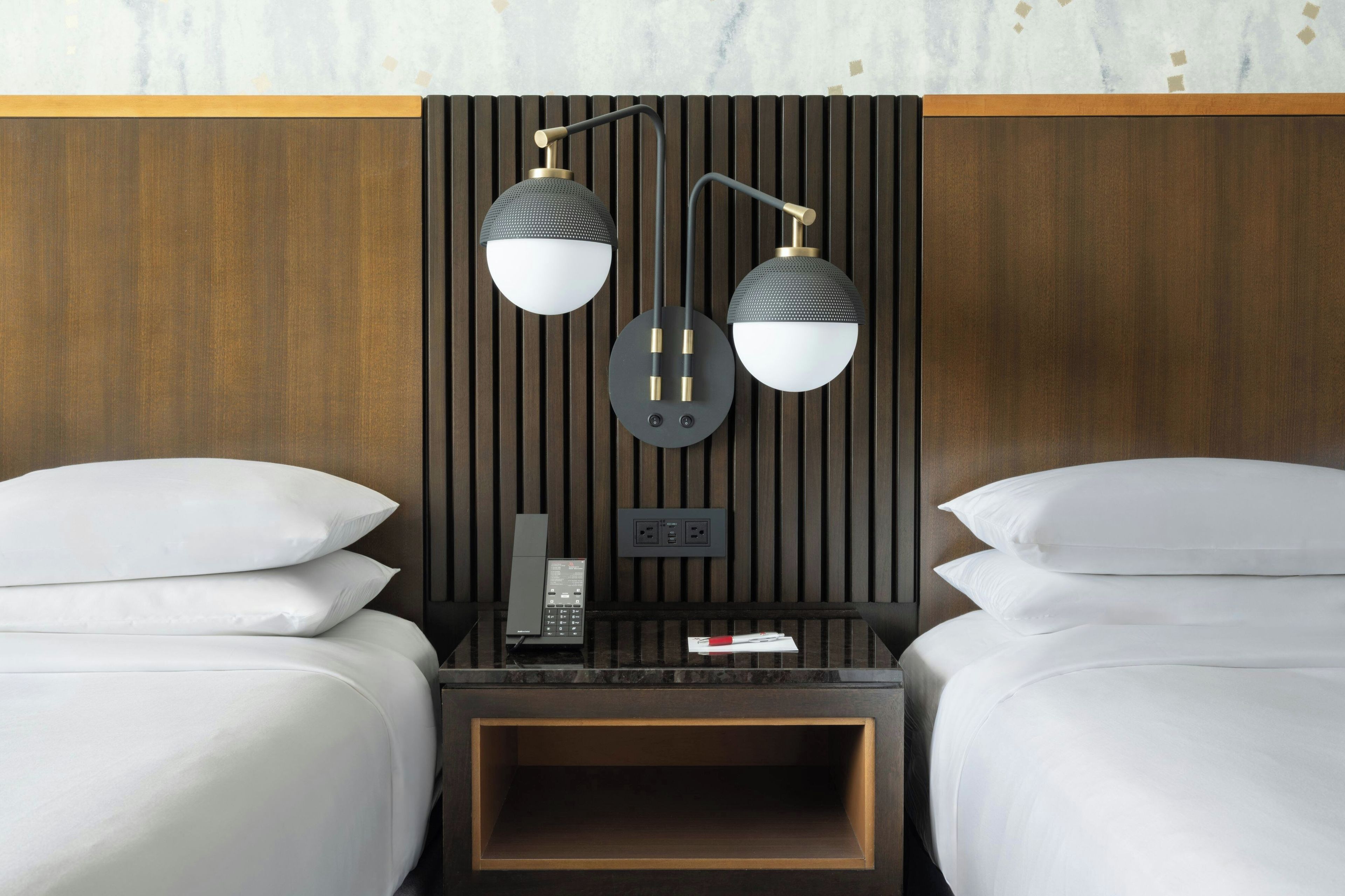 A hotel room with two beds, a nightstand with a phone and a remote, and a wall-mounted lamp.