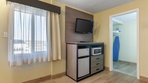 A hotel room with a wooden floor, a TV mounted on the wall, a minibar, and a window with sheer curtains. - SureStay by Best Western Chambers Petrified Forest (Chambers)
