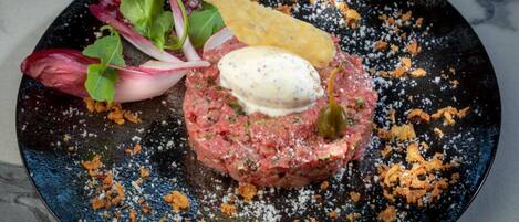 A steak tartare dish with a poached egg, garnished with herbs and a cherry, on a dark plate with scattered orange flakes.