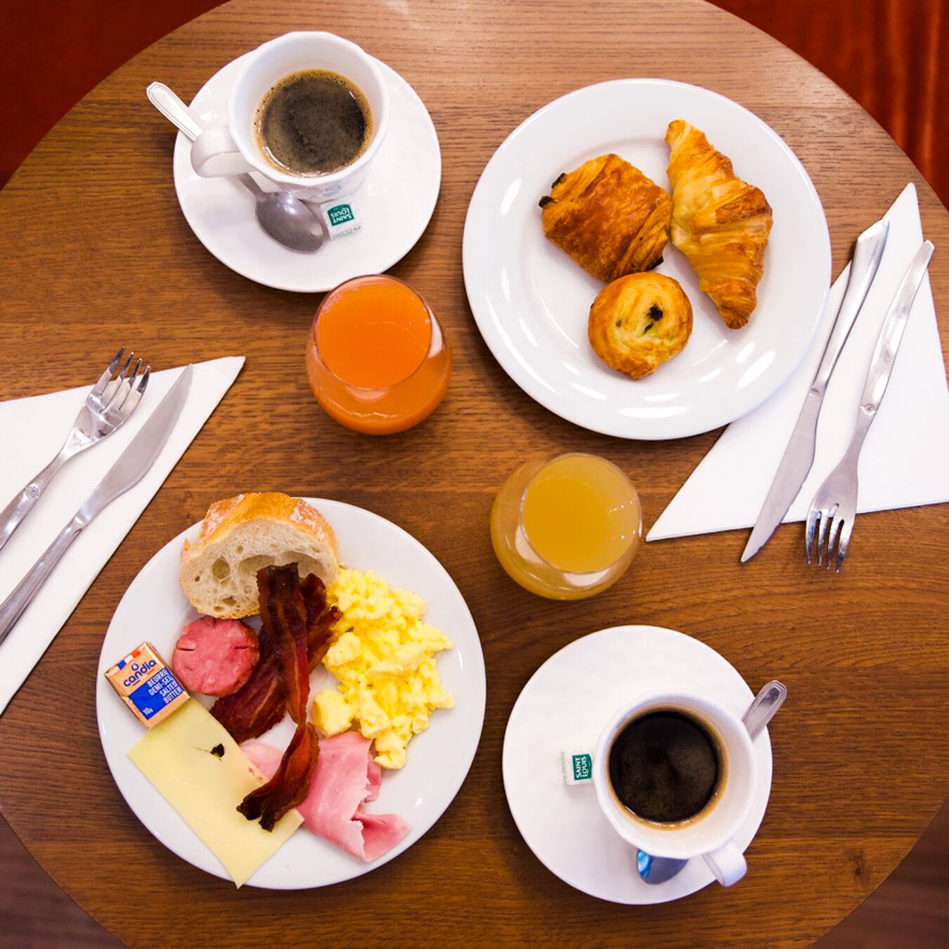 A breakfast table with plates of food, including eggs, bacon, ham, cheese, croissants, and juice. There are also two cups of coffee and a glass of orange juice.