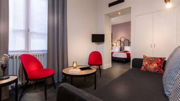 Une chambre d’hôtel moderne avec un canapé, deux fauteuils rouges, une petite table avec une lampe, un lit doté d’une tête de lit rouge et blanche, et un téléviseur fixé au mur.