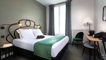 A hotel room with a bed, a desk, a chair, a lamp, and a window with curtains.