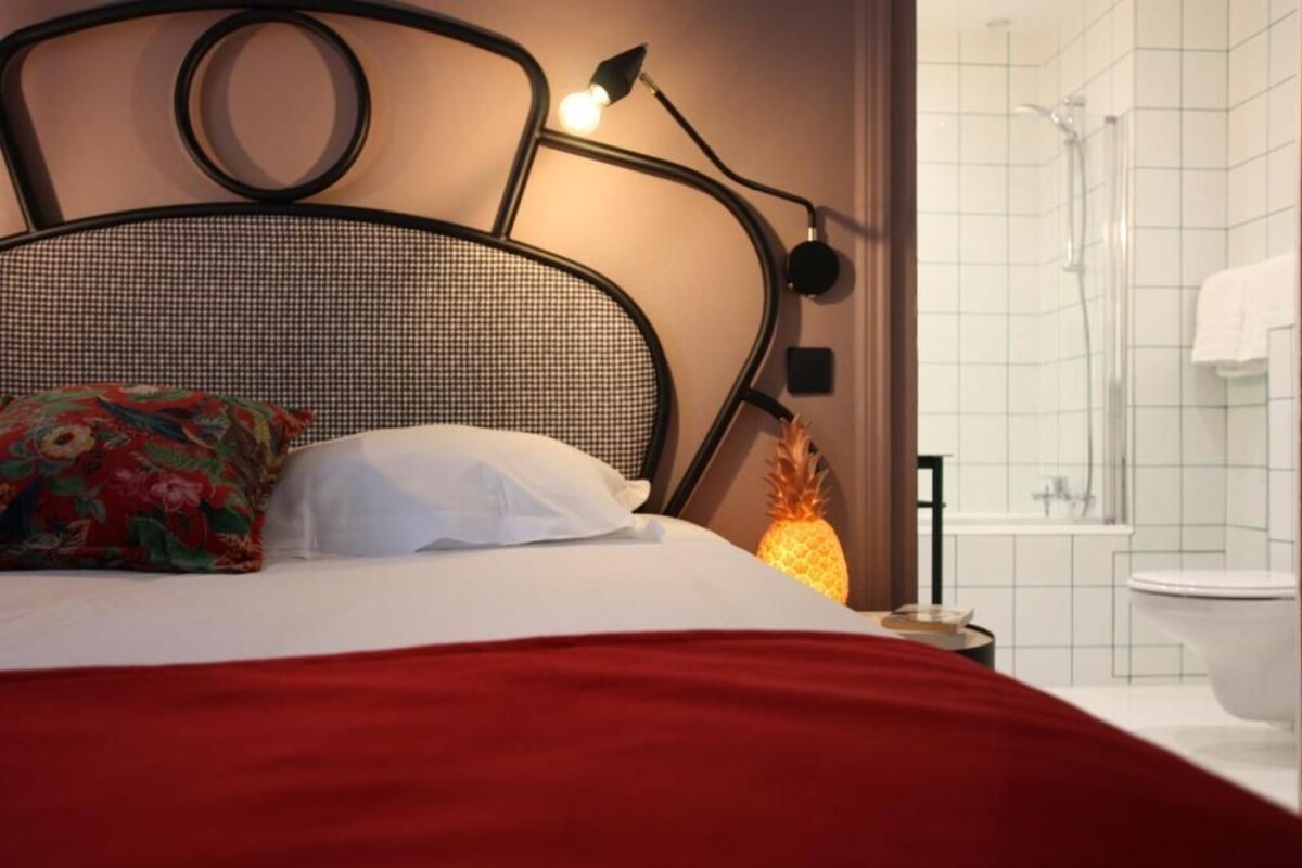 Una cama con una manta roja, una piña en la mesita de noche y una lámpara de pared.