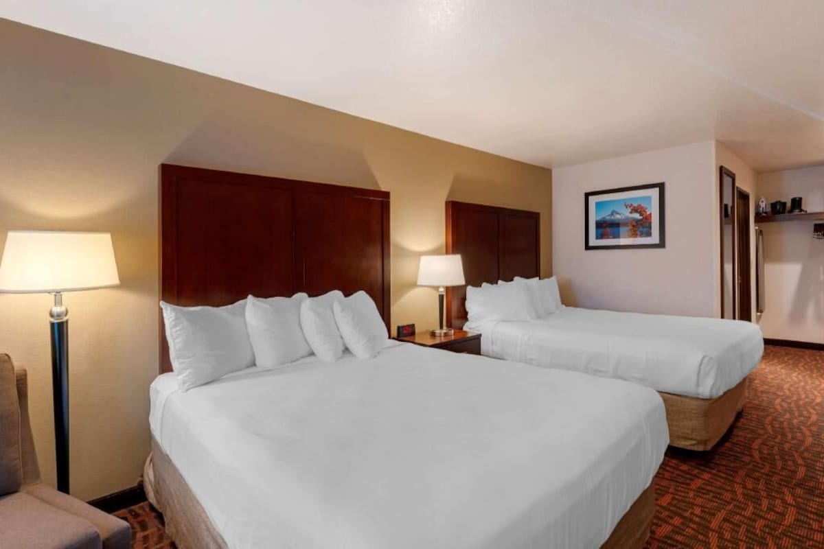 A hotel room with two beds, a desk, a chair, a lamp, a picture on the wall, and a small kitchenette.