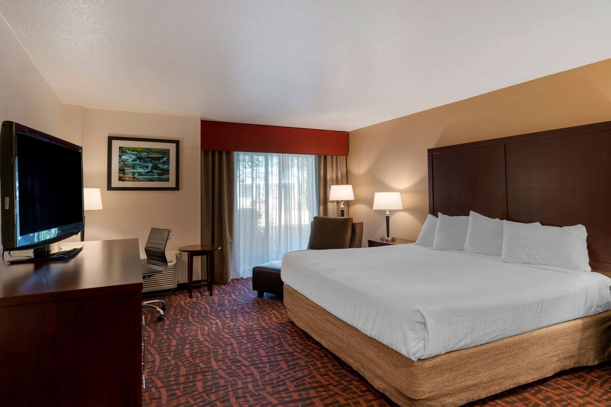 A hotel room with a large bed, a desk with a TV, a chair, a small table, and a window with curtains.