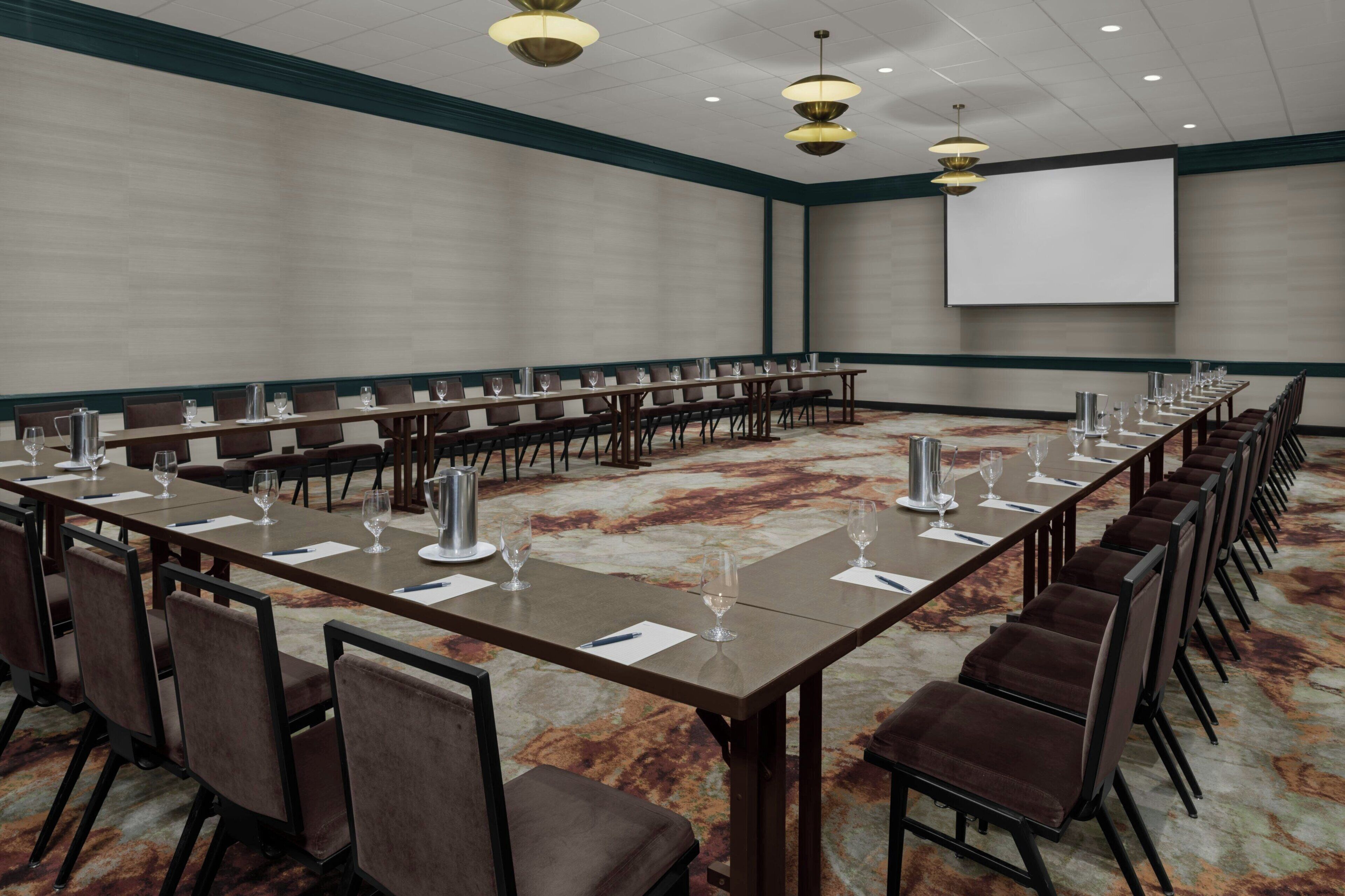 A conference room with a long table, chairs, a projector screen, and a carpeted floor.