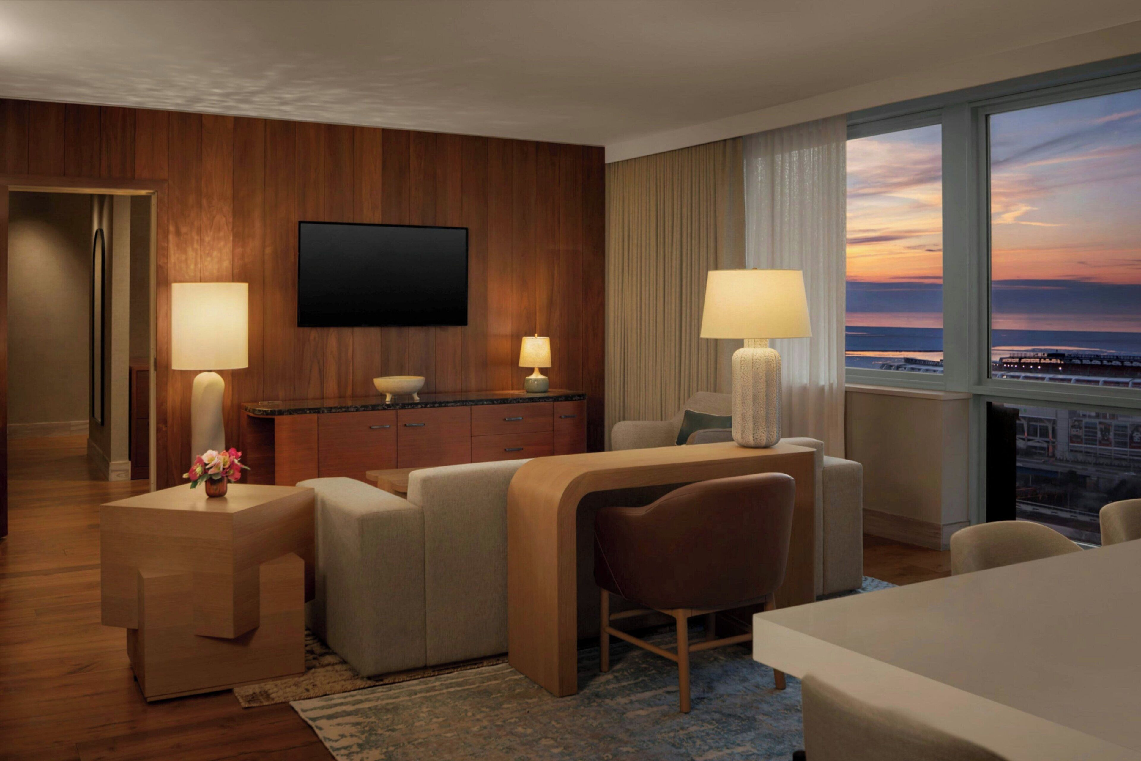 A hotel room with a flat-screen TV, a desk with two lamps, a sofa, a chair, and a view of the cityscape at sunset.
