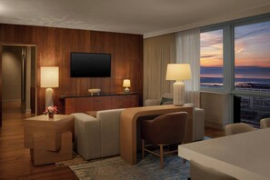 A hotel room with a flat-screen TV, a desk with two lamps, a sofa, a chair, and a view of the cityscape at sunset. - The Westin Cleveland Downtown (Cleveland)