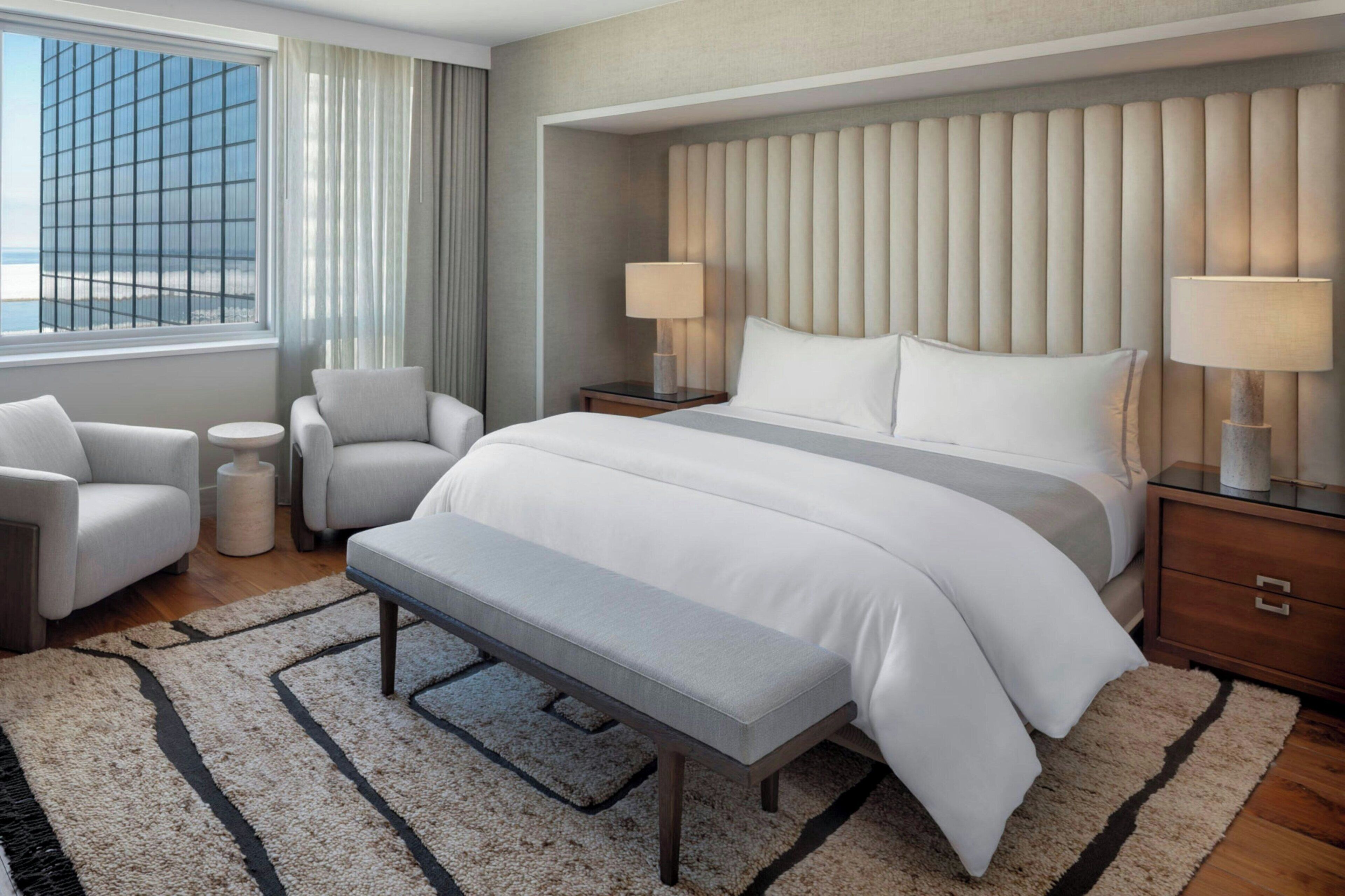 A hotel room with a large bed, two bedside tables with lamps, a bench, a small round table, and a view of a building through the window.