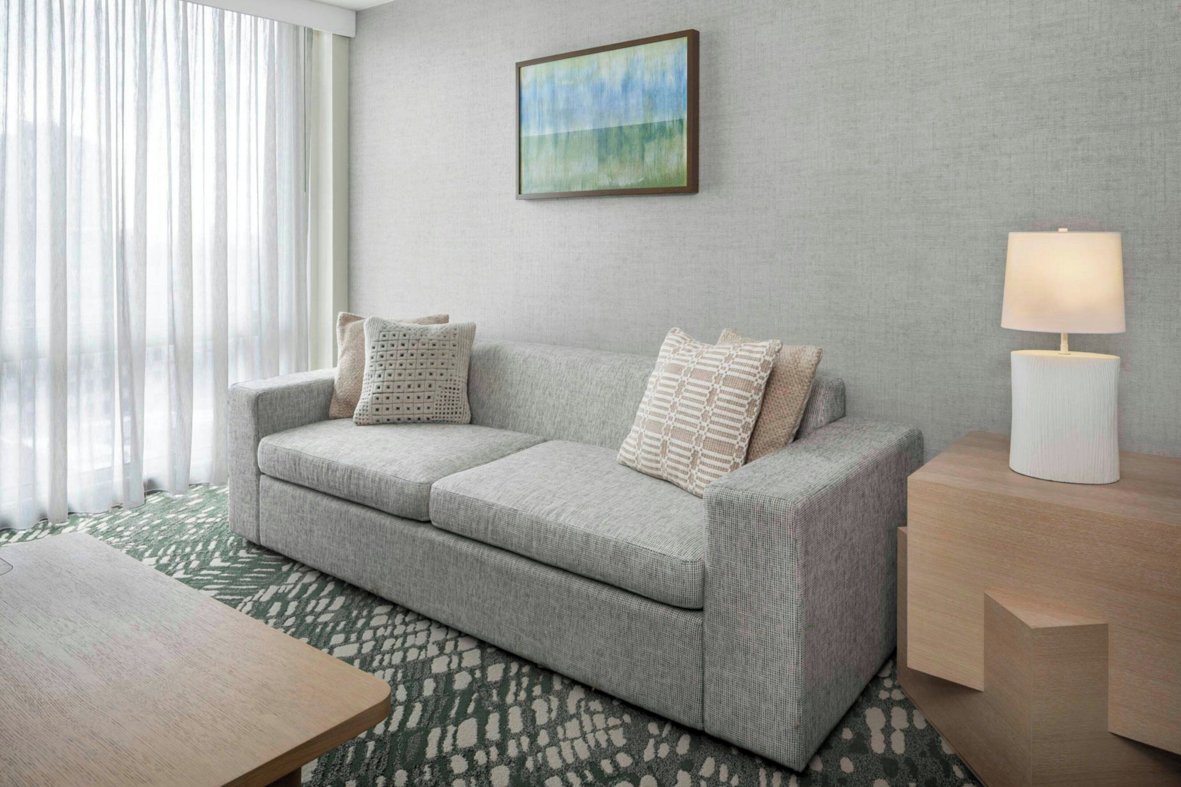 A modern living room with a grey sofa, patterned cushions, a painting on the wall, and a lamp on a side table.