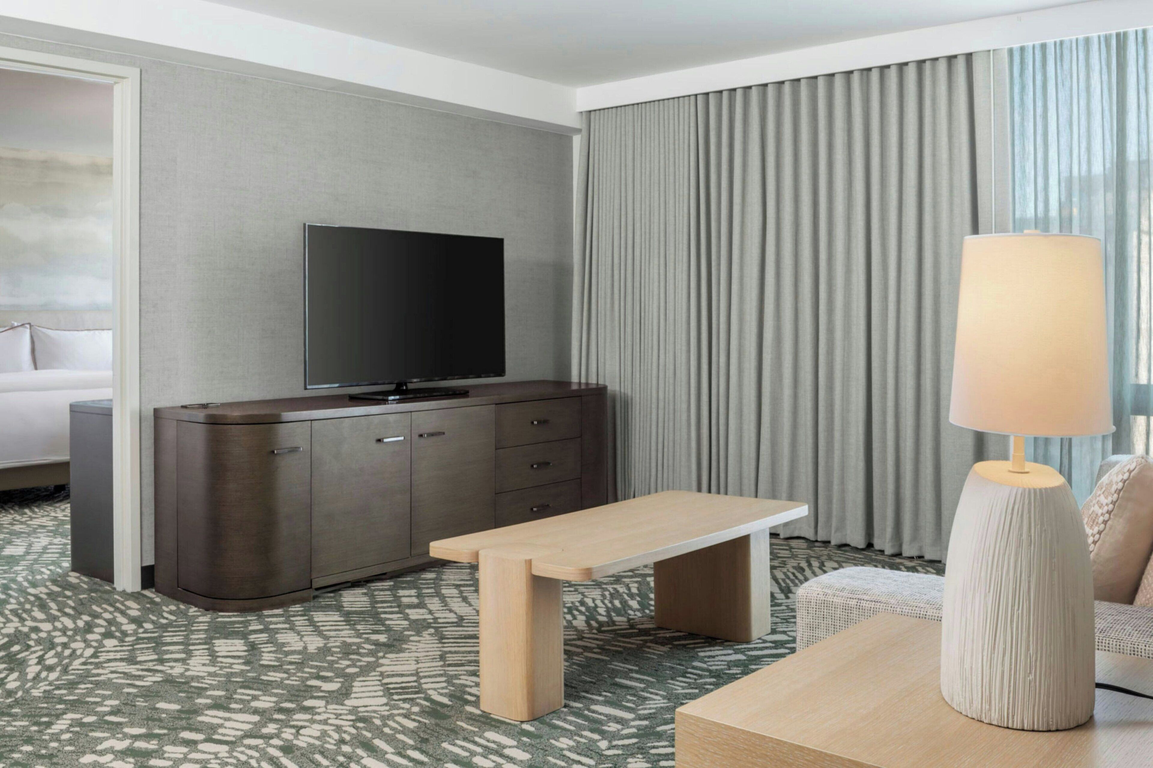 Habitación de hotel con televisión de pantalla plana sobre un mueble oscuro, un banco de madera, una lámpara y una alfombra estampada.