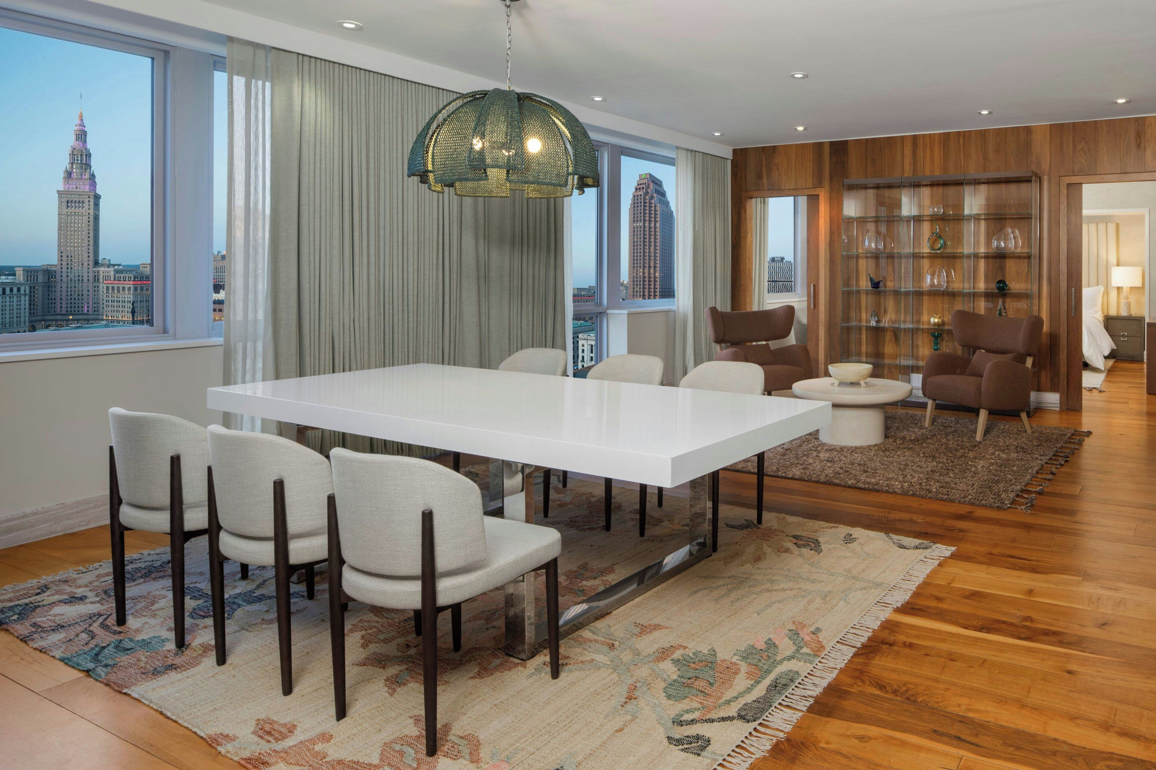 A modern living room with a dining area, large windows, a city view, a bookshelf, and a unique hanging light fixture.