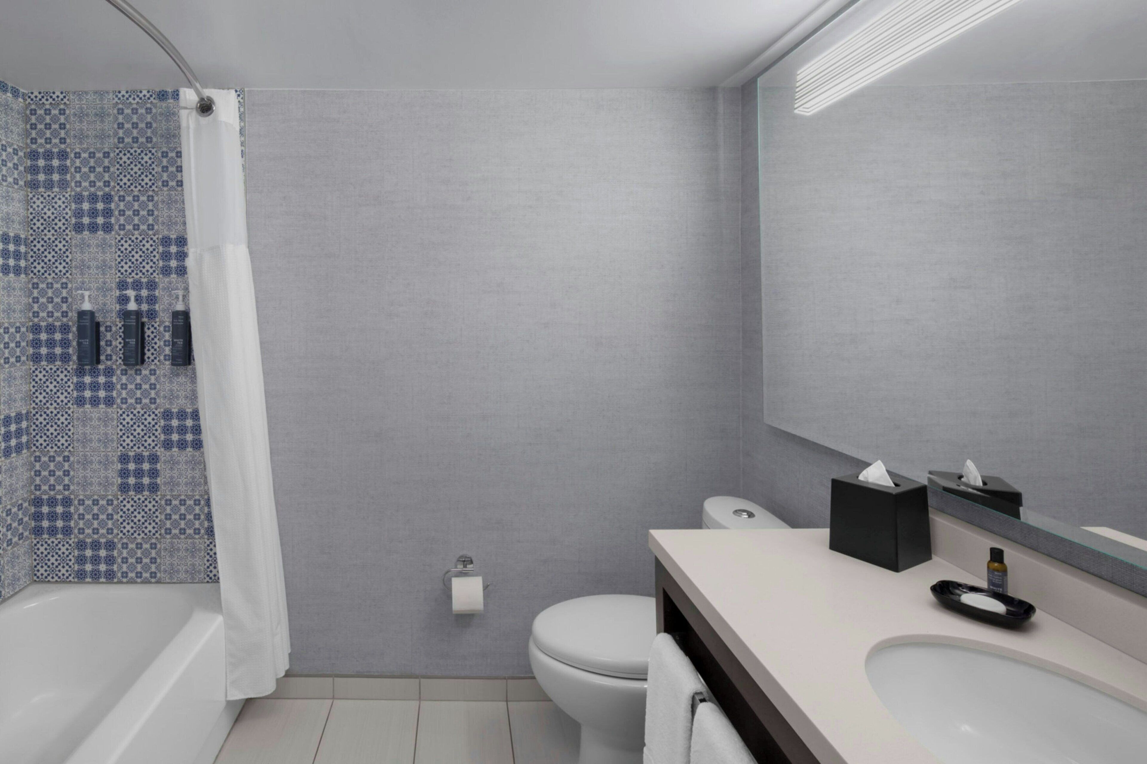 A modern bathroom with a grey tiled wall, a white toilet, a bathtub, and a vanity with a mirror.