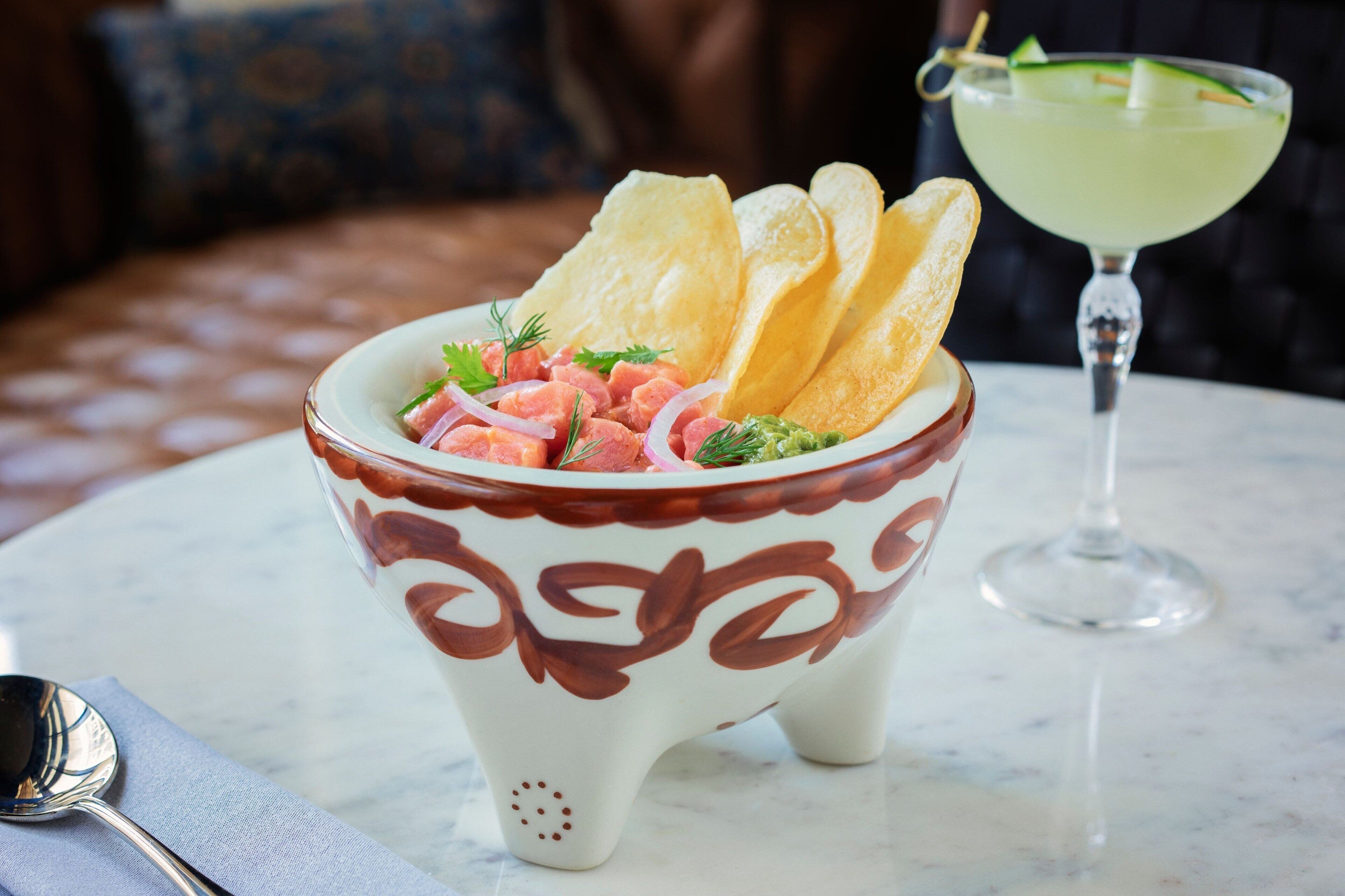 A bowl of ceviche with chips and a lime margarita on a table.