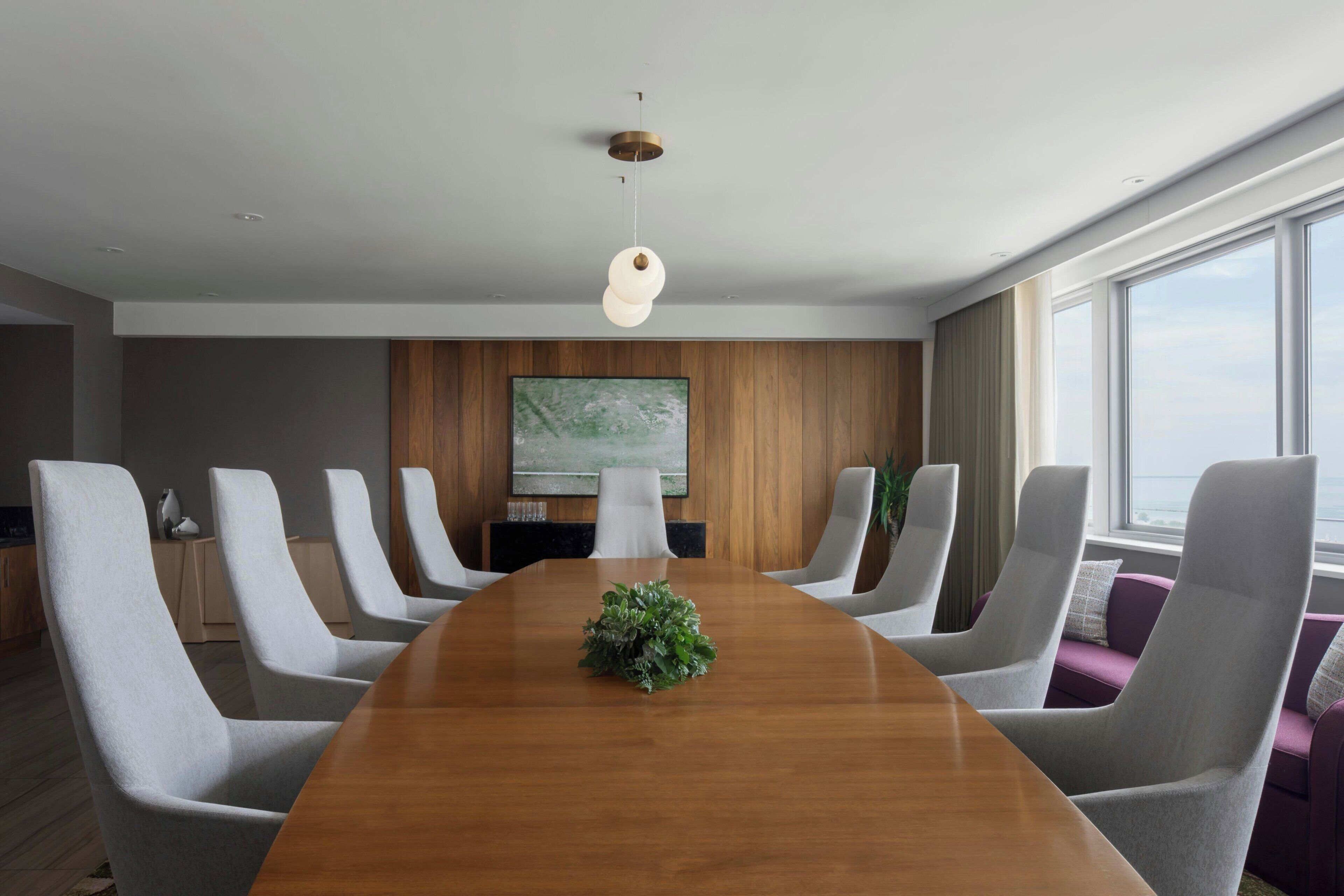 Een moderne vergaderruimte met een lange houten tafel, zes grijze gestoffeerde stoelen, een grote flatscreen televisie aan de muur en een raam met uitzicht.