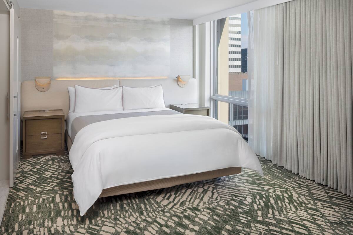A hotel room with a bed, nightstands, a wall-mounted light, a window with curtains, and a patterned carpet.