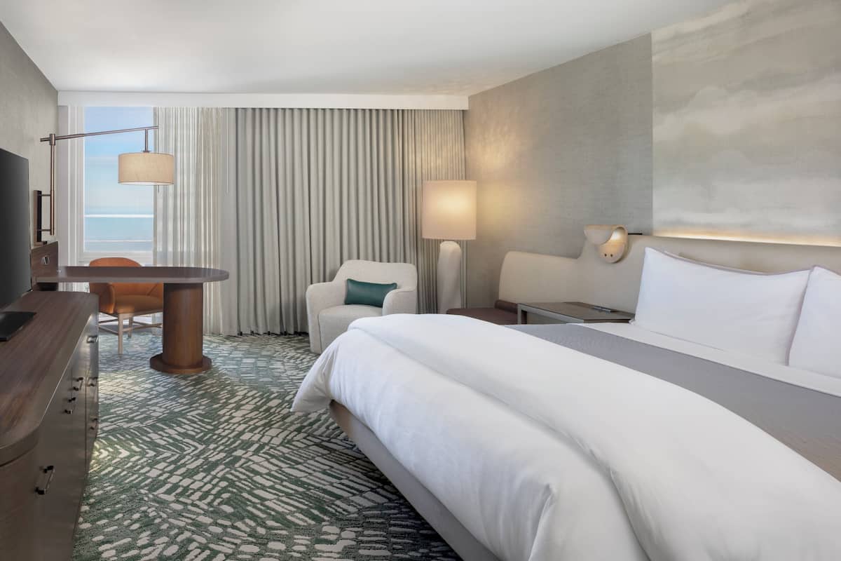 A hotel room with a bed, a desk, a chair, a lamp, and a view of the ocean.