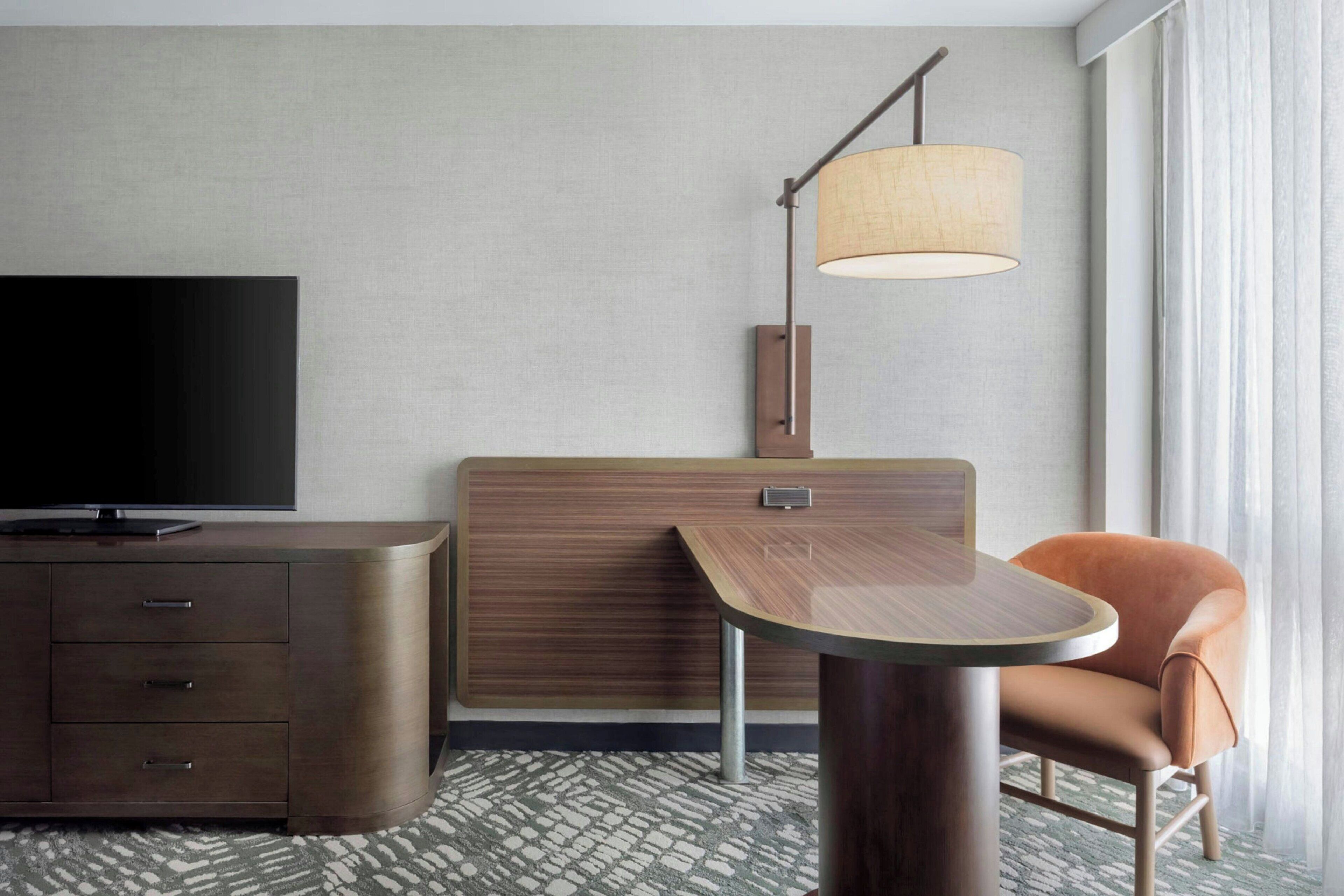 A hotel room with a flat-screen TV, a wooden desk, a chair, and a lamp.