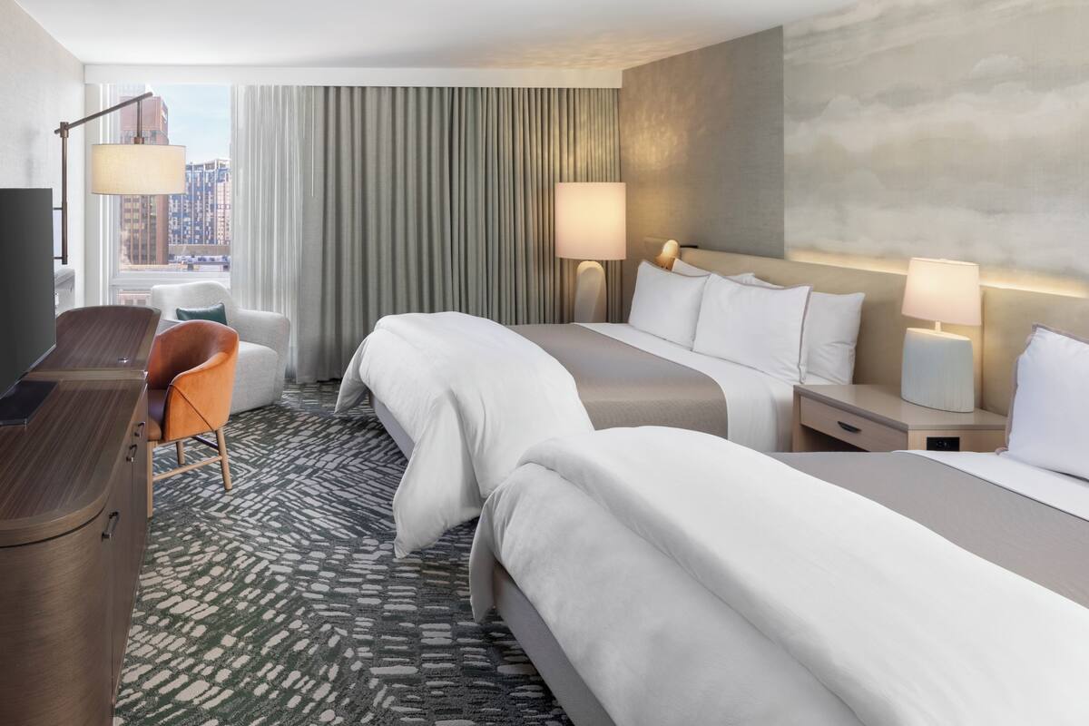 A hotel room with a bed, two bedside tables, a desk, a chair, and a large window with curtains.
