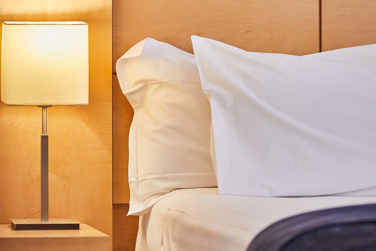 A neatly made bed with white linens and pillows, a bedside table with a lamp, and a wooden headboard.