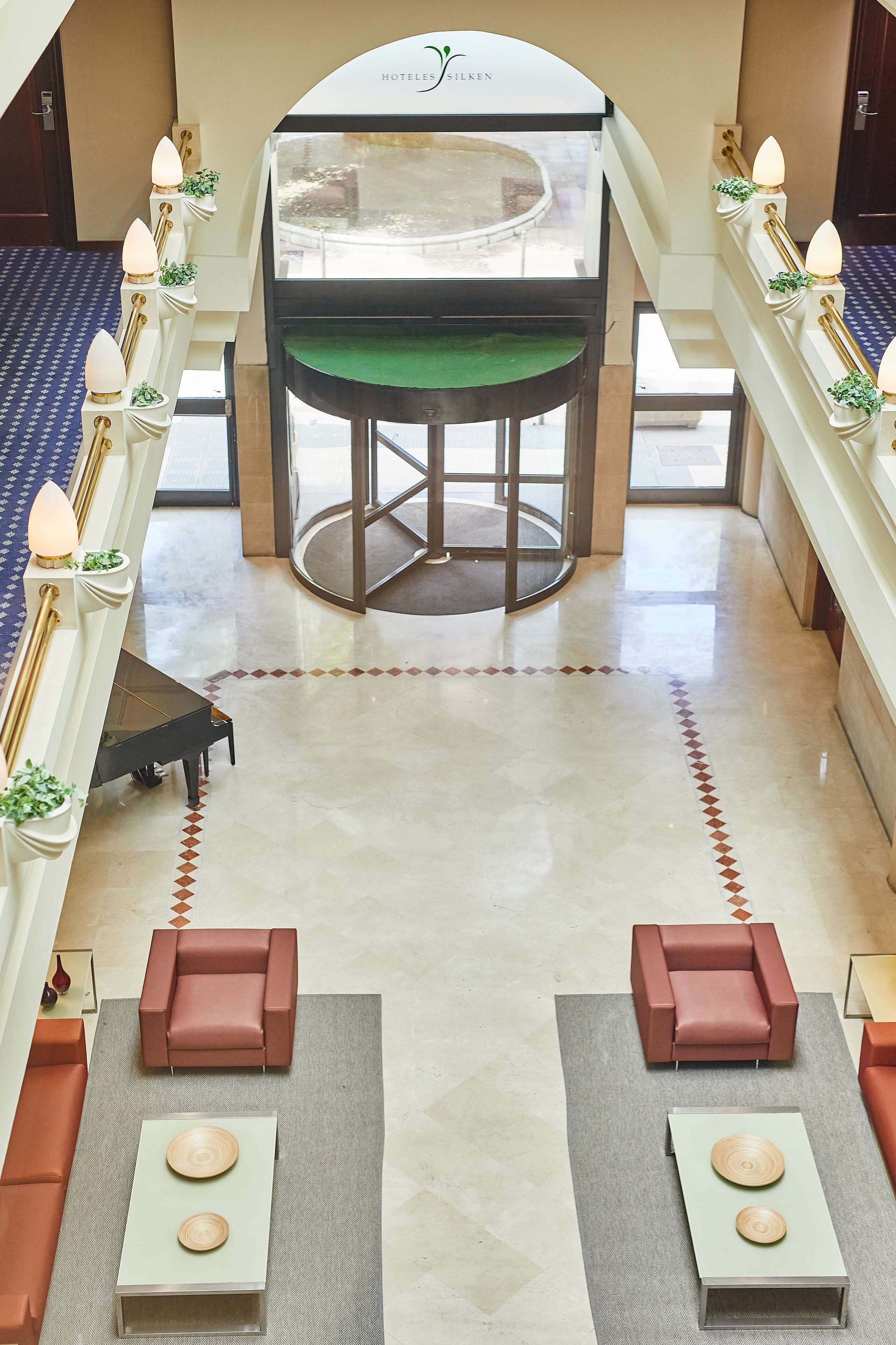 Un vestíbulo de hotel con un ascensor central, dos sofás, un piano y una mesa de recepción.