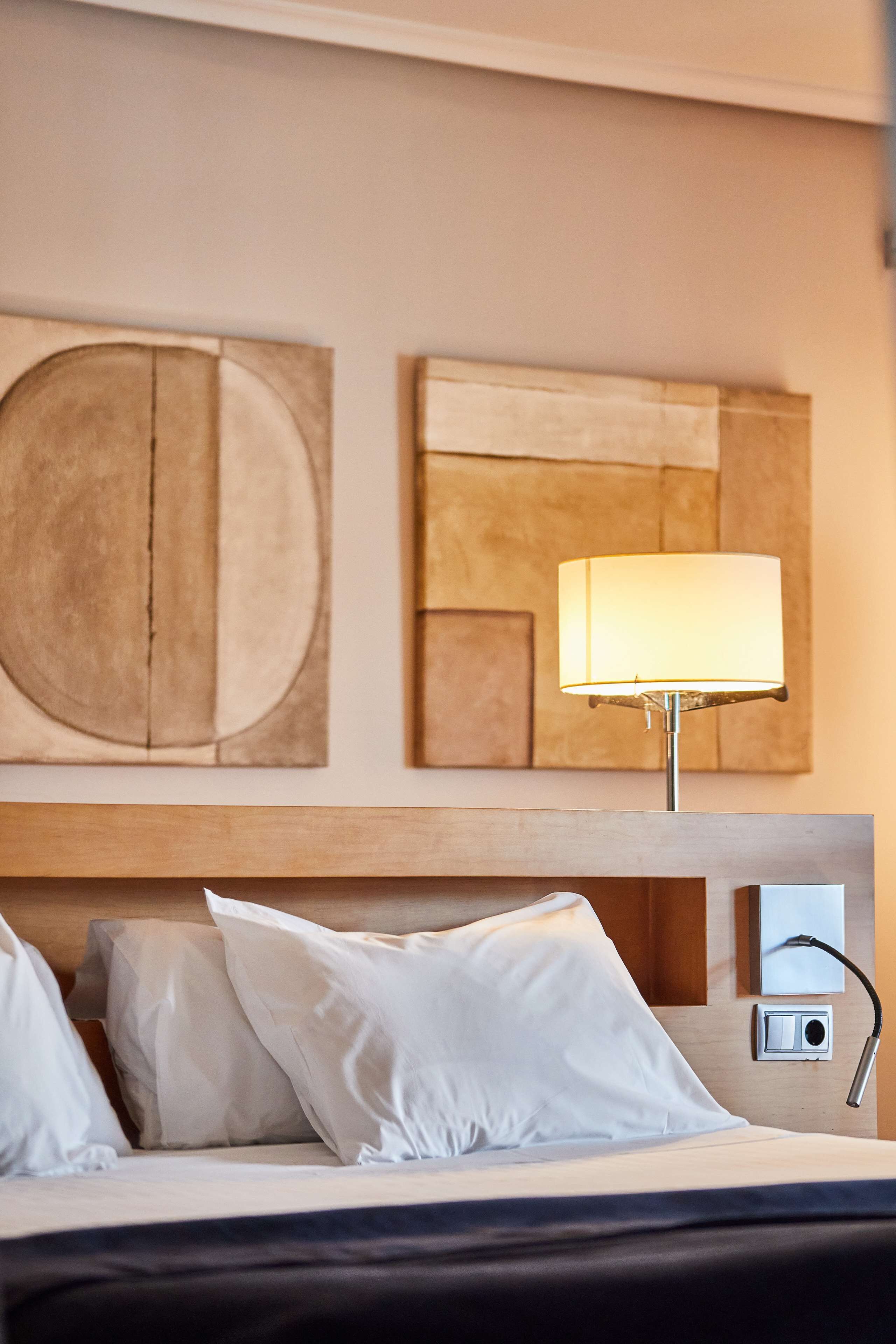 Een hotelkamer met een bed, witte kussens, een lamp en abstracte muurkunst.