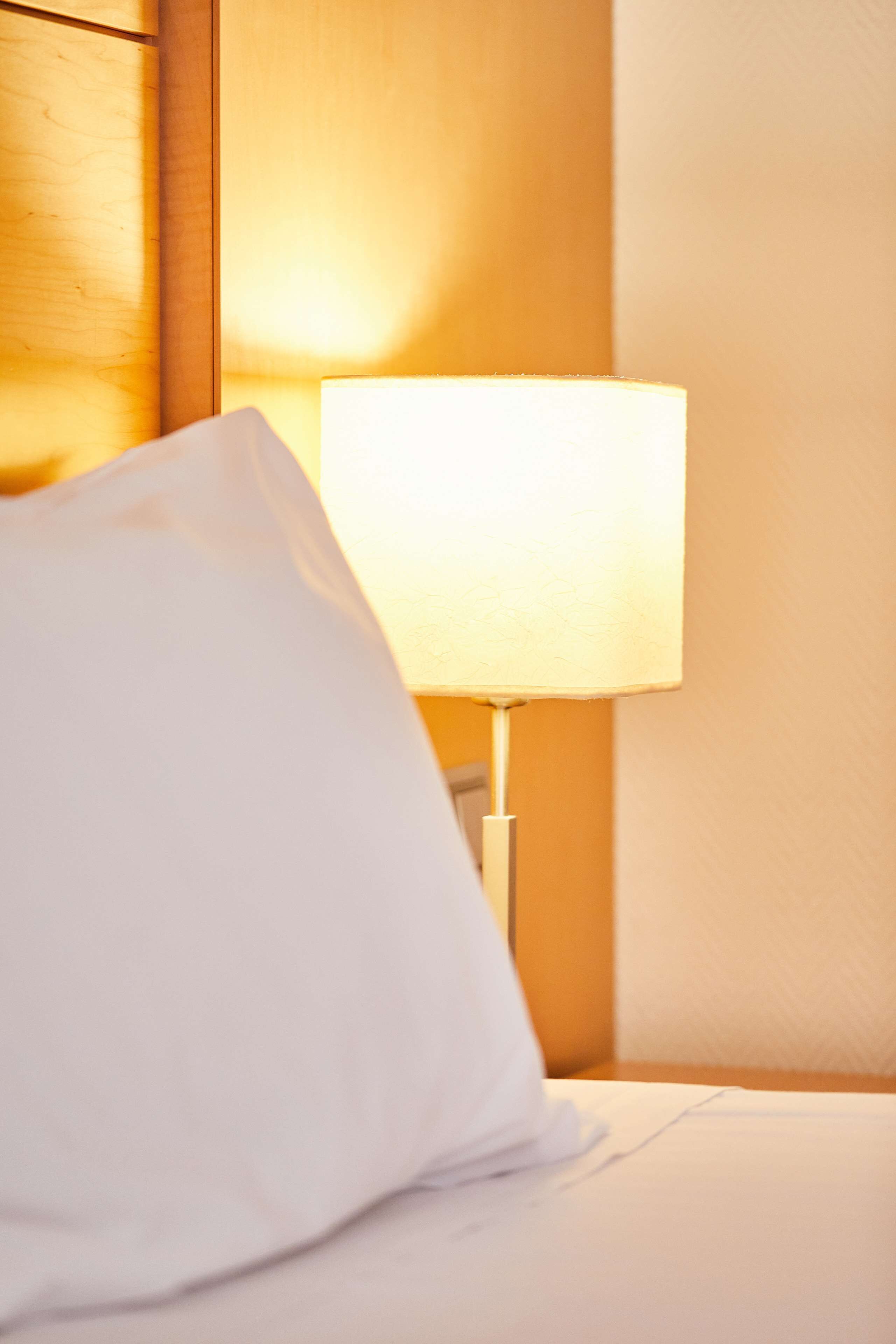 A hotel room with a bed, a white pillow, and a bedside lamp.