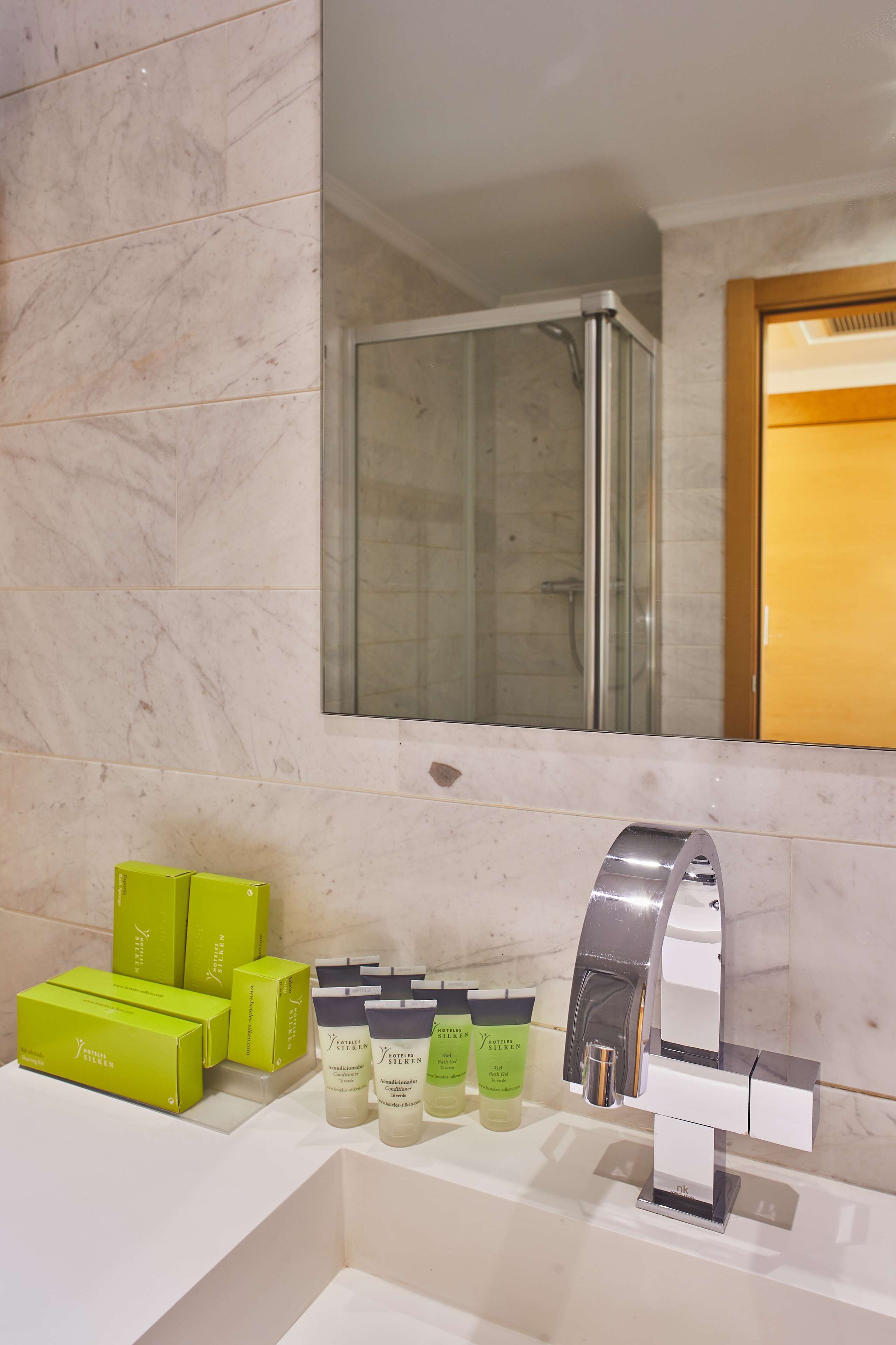 Un baño con pared de mármol, cabina de ducha de vidrio y lavamanos con distintos artículos de aseo.
