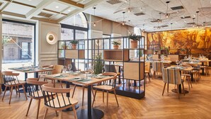 A modern restaurant with wooden flooring, multiple dining tables, and chairs. There are potted plants on shelves and a large mural on the back wall. - Hotel Silken Ciudad de Vitoria (Vitoria-Gasteiz)
