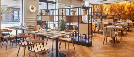 Un restaurante moderno con suelo de madera, varias mesas y sillas para comer. Hay plantas en macetas sobre estantes y un mural grande en la pared de fondo.