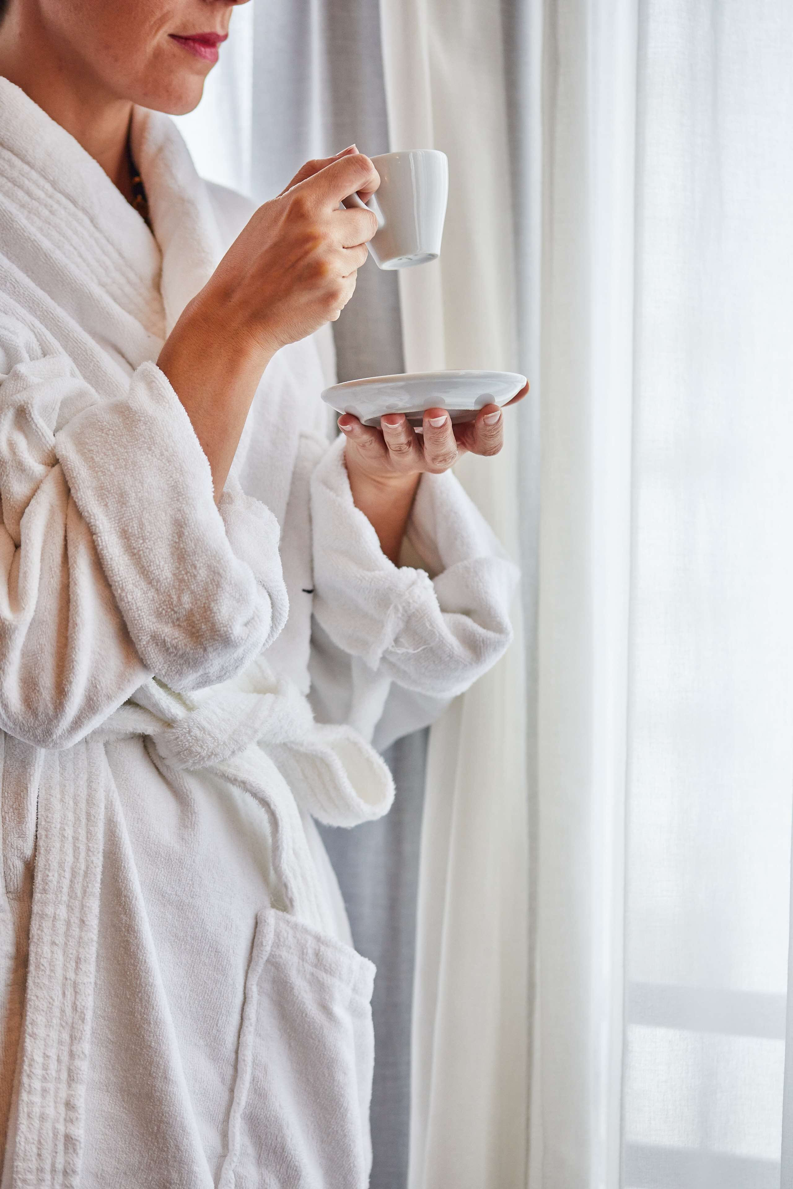 A person in a white bathrobe holding a cup and saucer, standing near a window with curtains.