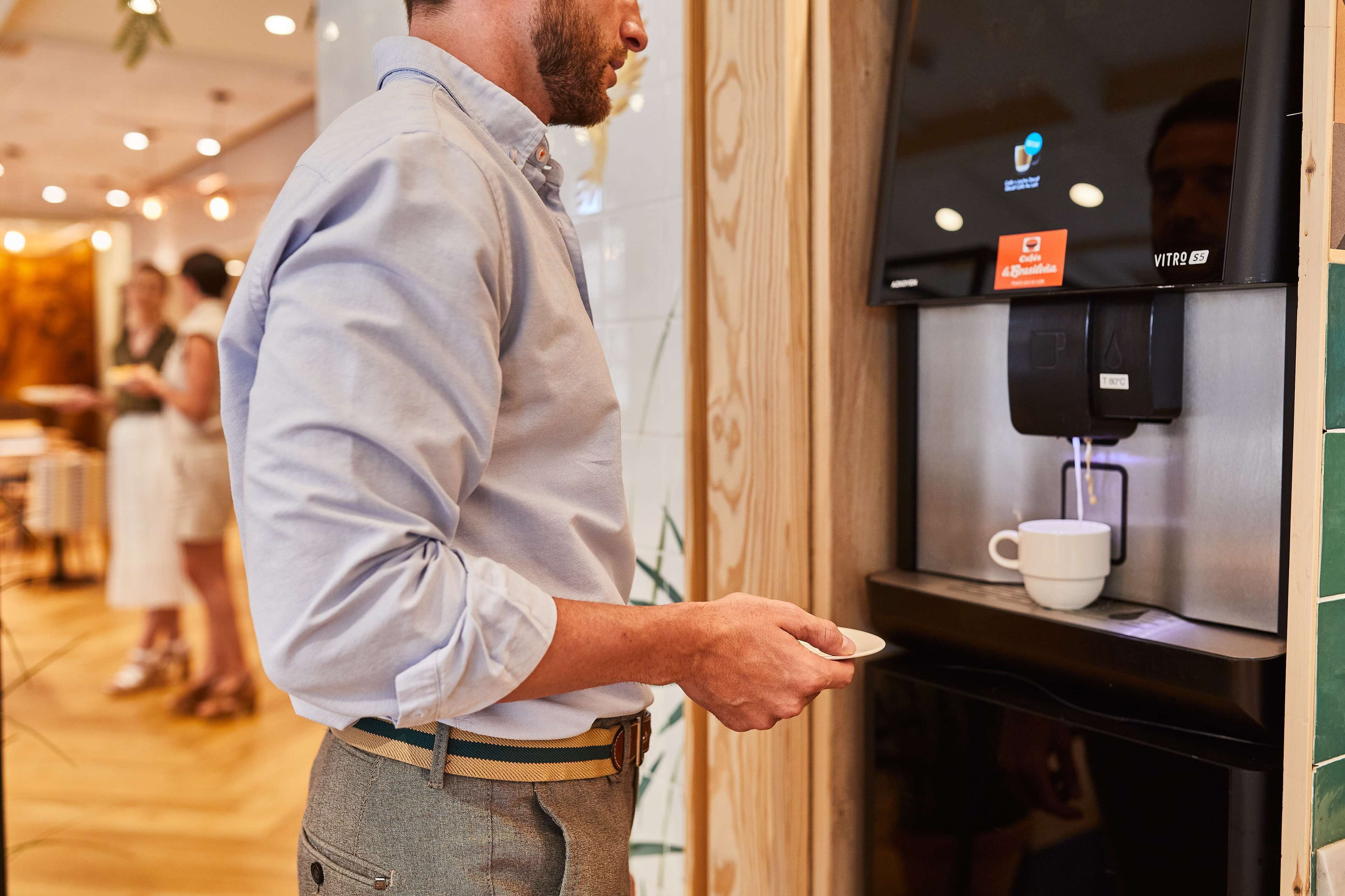 A person using a coffee machine in a public area with a wooden counter and a digital display.