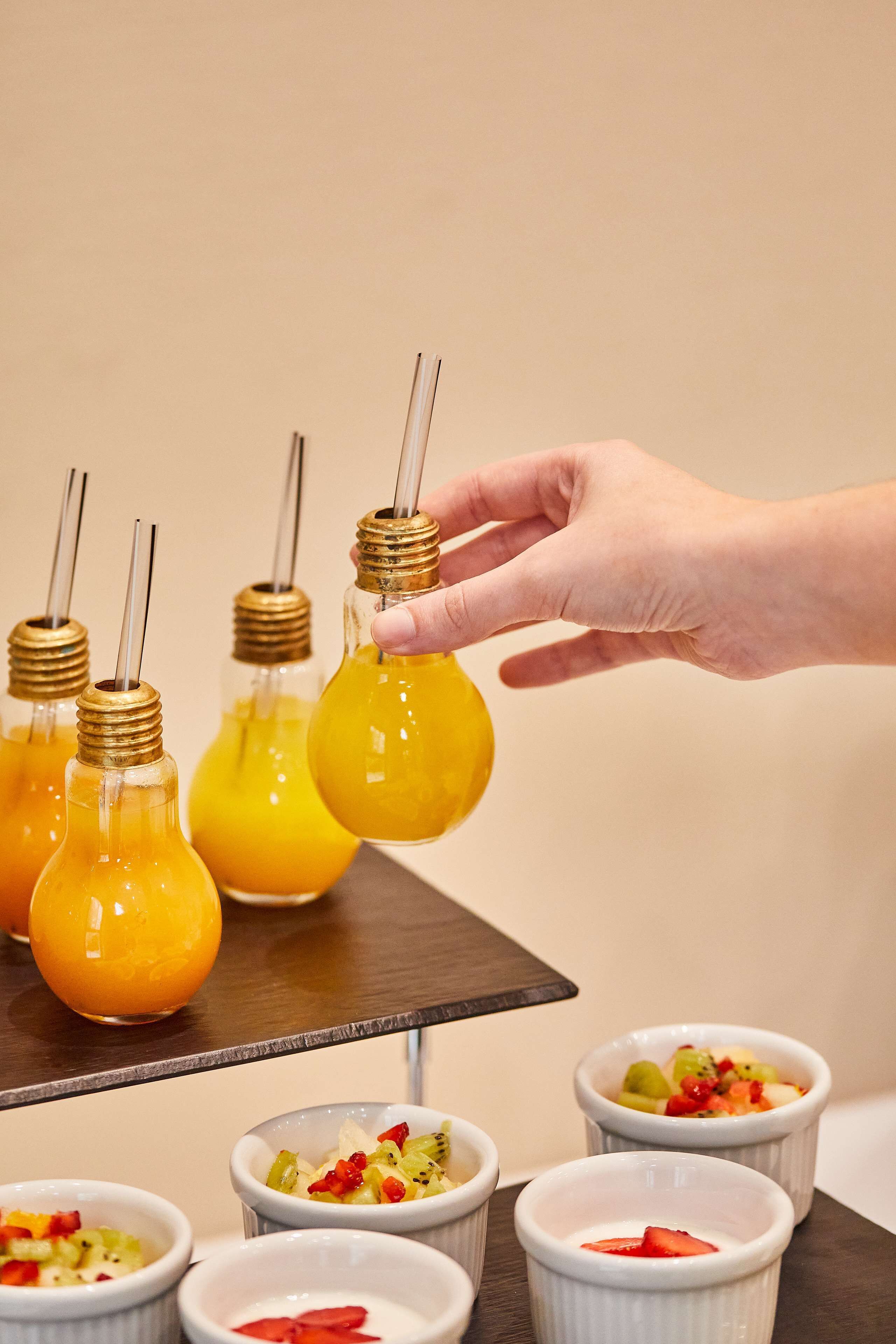 A hand reaching for a light bulb-shaped glass with a yellow liquid, surrounded by similar glasses and small bowls with fruit toppings.