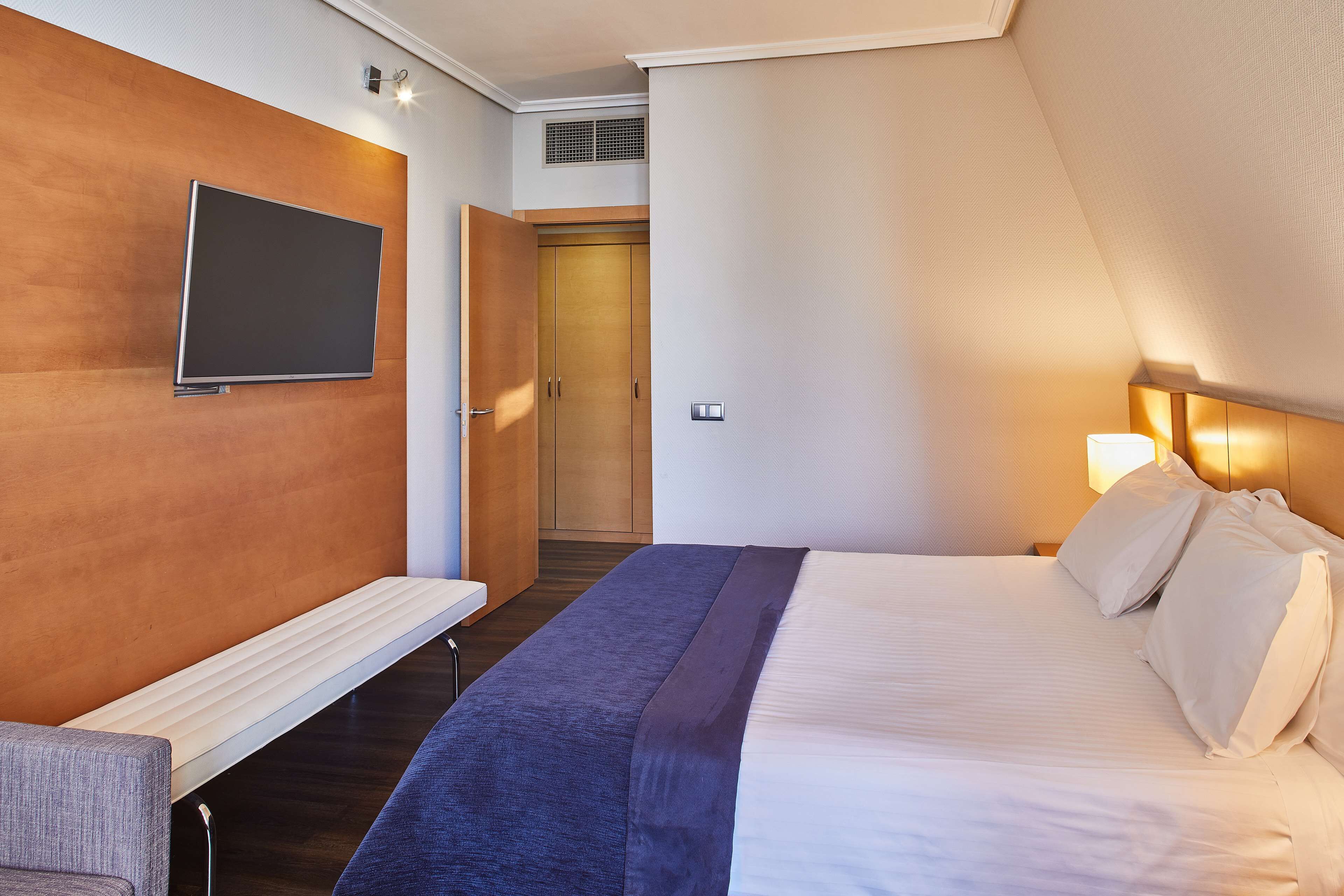 Een hotelkamer met een bed, een aan de muur bevestigde televisie, een bankje en een deur naar een andere kamer.