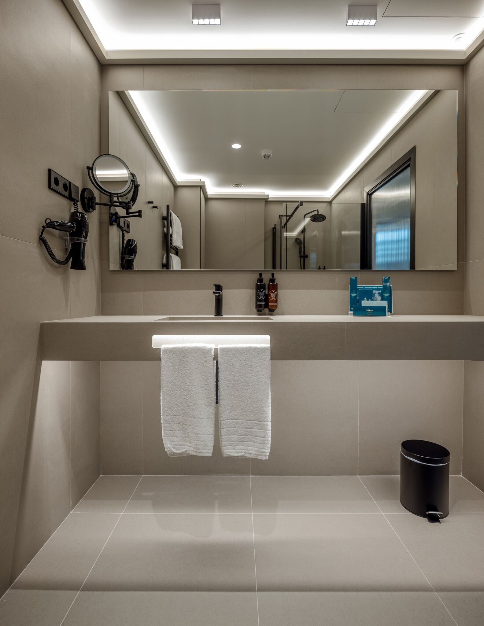 A modern bathroom with a large mirror, a towel rack with two towels, a sink, and a black trash can.
