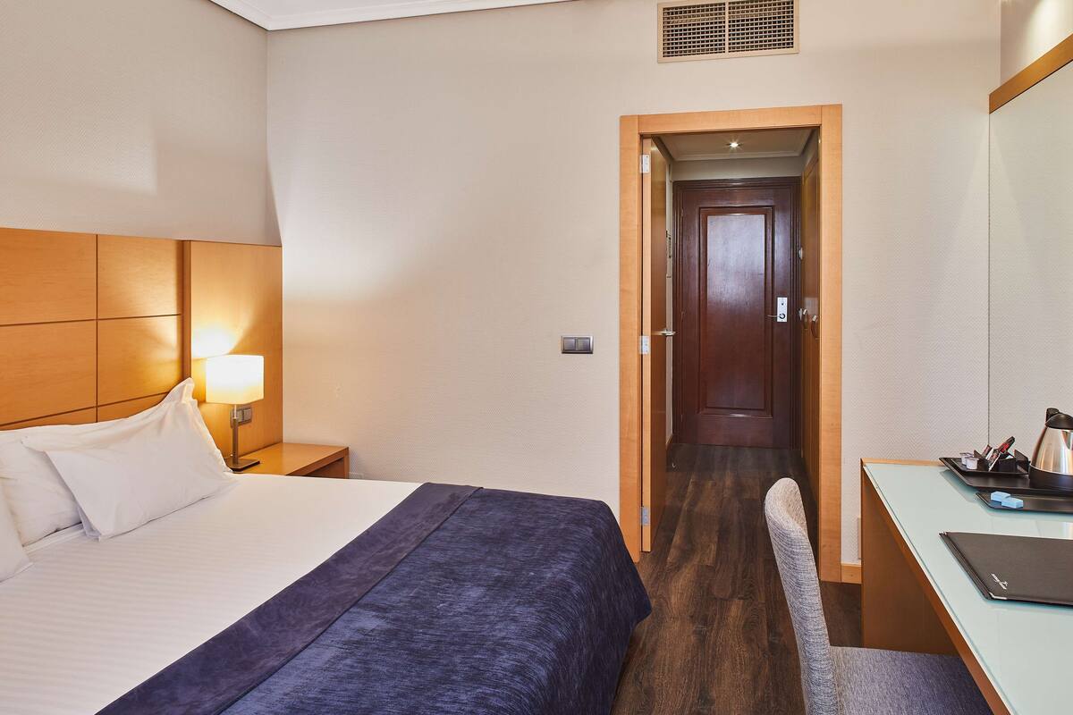 A hotel room with a bed, bedside table, lamp, and a desk with a chair.