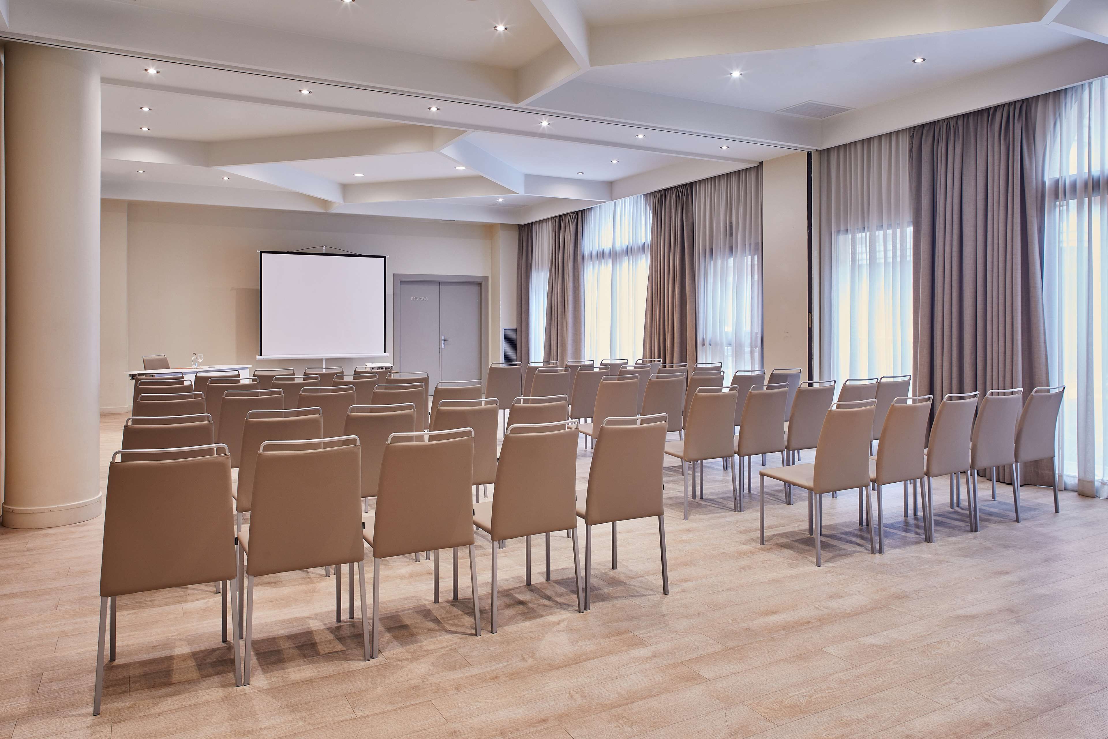 Un salón de conferencias con filas de sillas, una pantalla de proyector y ventanas grandes con cortinas.