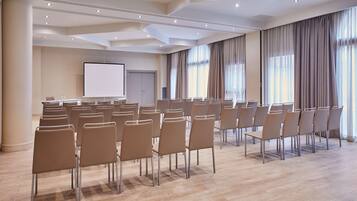 Un salón de conferencias con filas de sillas, una pantalla de proyector y ventanas grandes con cortinas.