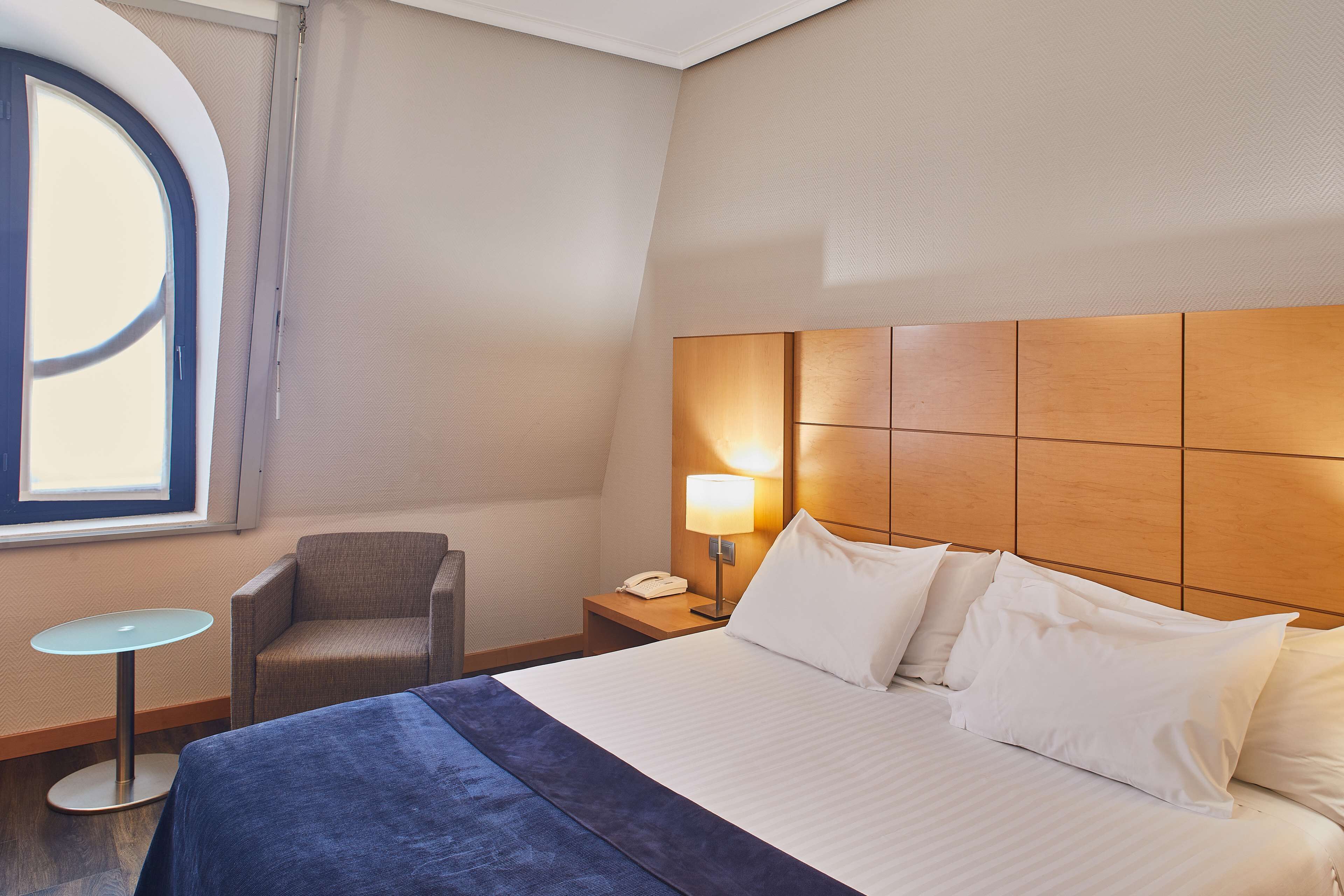 Habitación de hotel con cama, una silla, una mesita y una mesita de noche con lámpara.