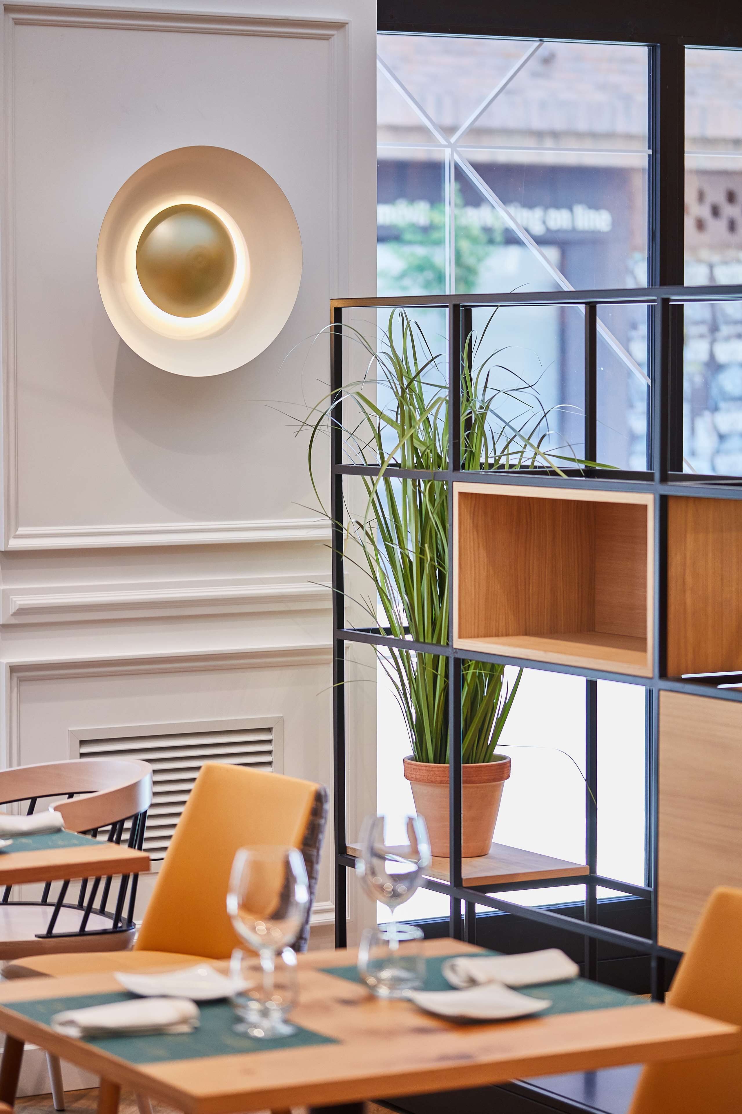 A restaurant interior with a modern design, featuring a wall-mounted light fixture, wooden shelves, and a potted plant.