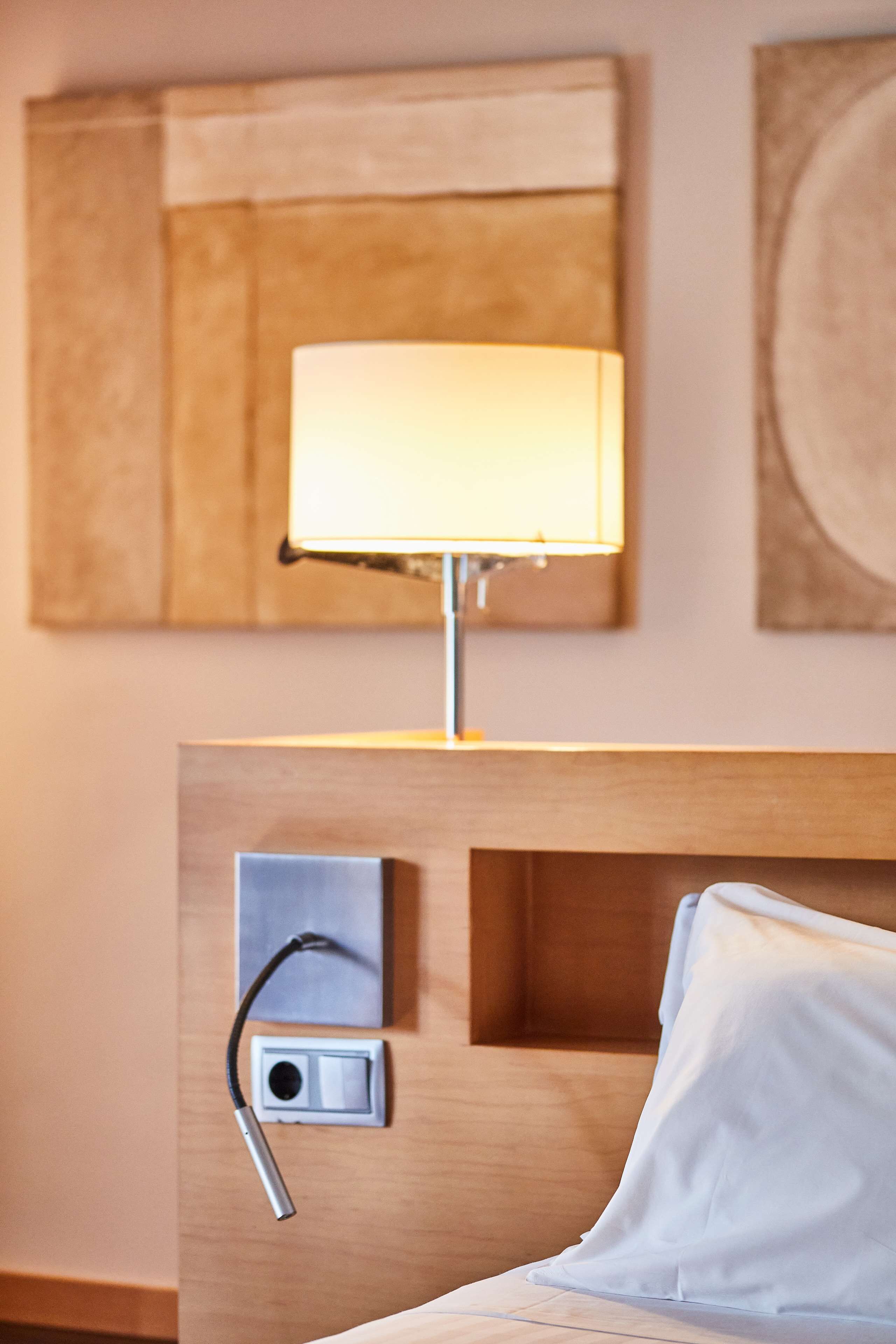 Habitación de hotel con cama, lámpara de noche, toma de corriente y una obra de arte colgada en la pared.
