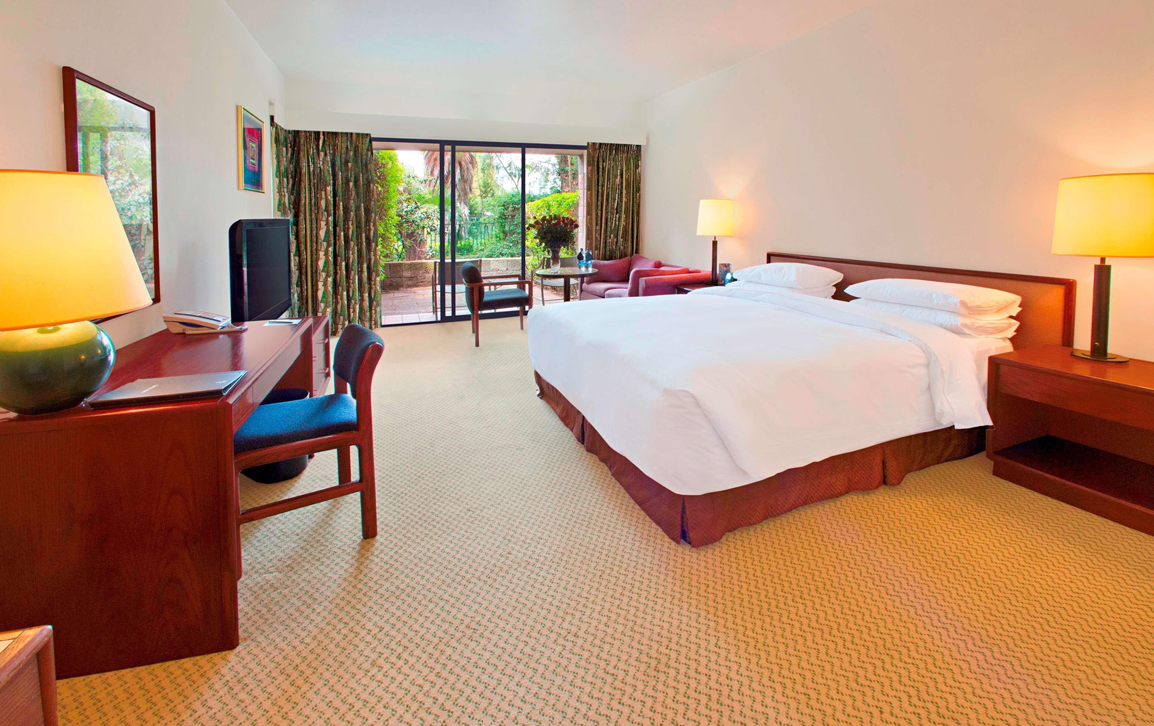 A hotel room with a bed, two bedside tables with lamps, a desk with a chair, a television, and a view of the outdoors through a glass door.